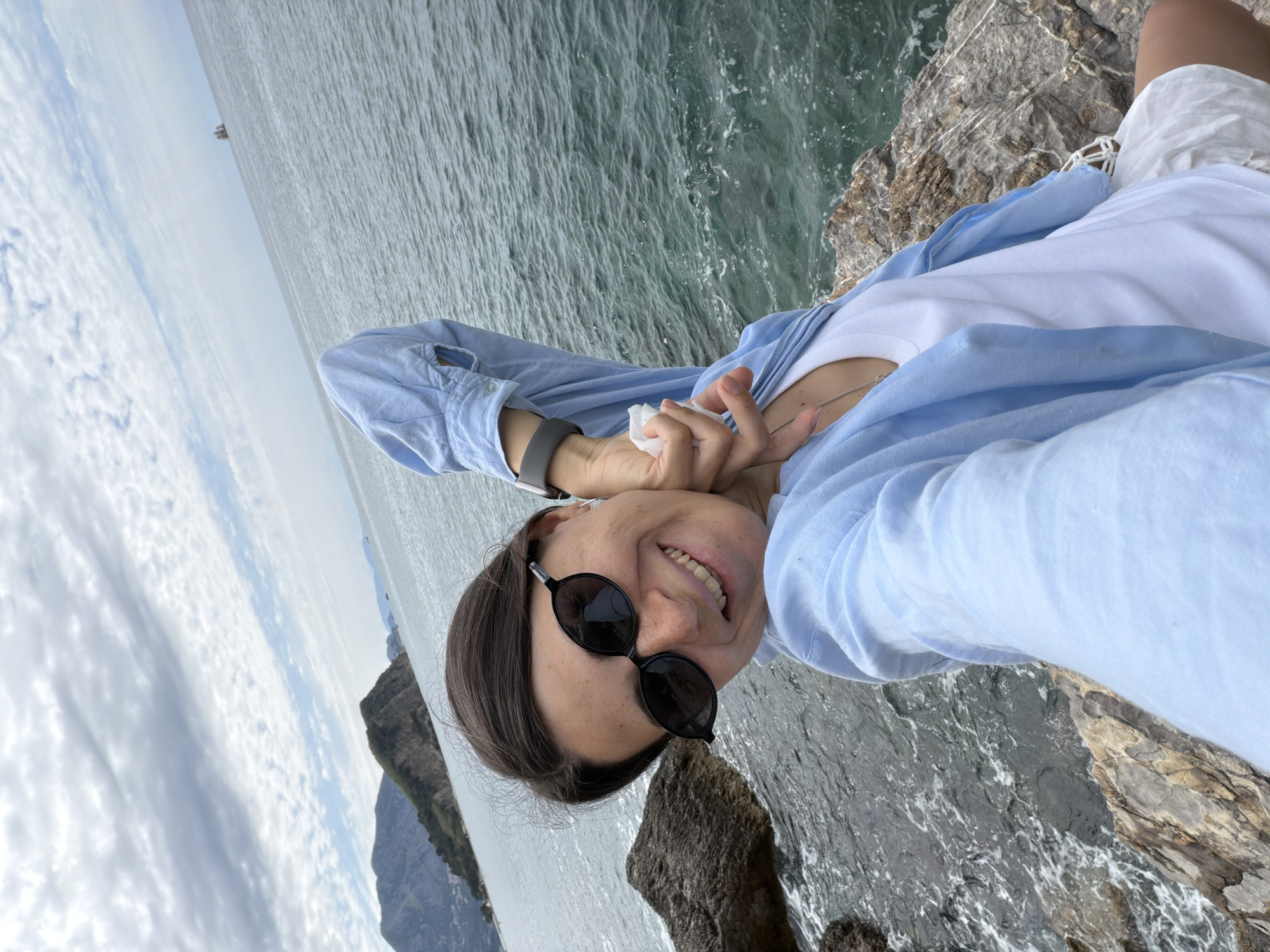 A person, presumably female, takes a selfie outdoors. The subject is wearing sunglasses, a white t-shirt under a light blue shirt, and a grey wristwatch on the left wrist. They are standing near a body of water, potentially the sea, with rocky terrain below and to the right. There's a mountain in the background on the left. The sky is partially covered with clouds. Given the location information, this is likely along the coastline near Budva, Montenegro. No specific actions or interactions, except the selfie being taken, are evident.
