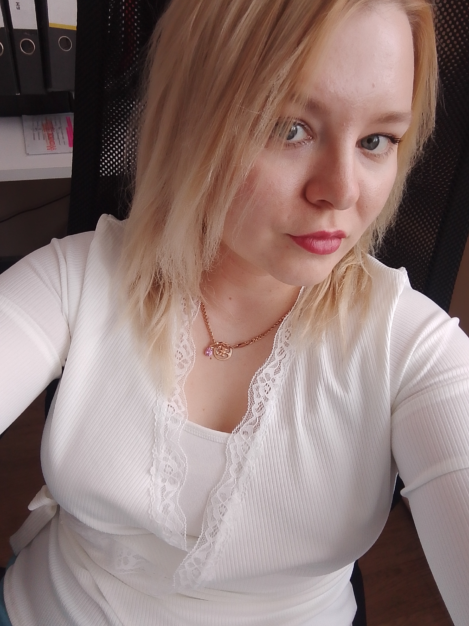 A blonde-haired woman is looking directly at the camera. She is wearing a white, long-sleeved shirt with lace detailing at the neckline, and a gold necklace with a charm and a small pink gem. She has fair skin, and is wearing lipstick. The background shows black office chair mesh, binders on a shelf, and a desk surface. The location is unknown and there is no indication of being located in the city of Izhevsk, Russia.