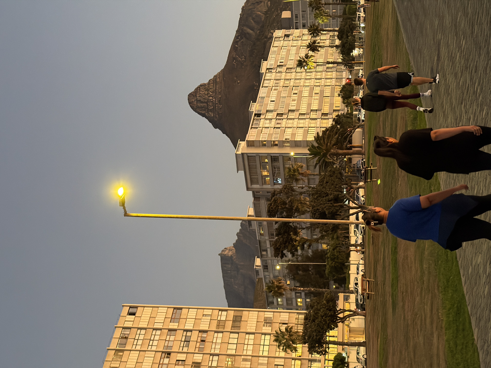 The image depicts a promenade in Cape Town, South Africa, at dusk. In the background are Lion's Head mountain and a building complex with windows. A yellow street lamp illuminates the foreground. Three people are walking along the promenade; one in a blue shirt, one in black, and another in gray shorts and a black shirt. Cars and palm trees are also visible in the background.