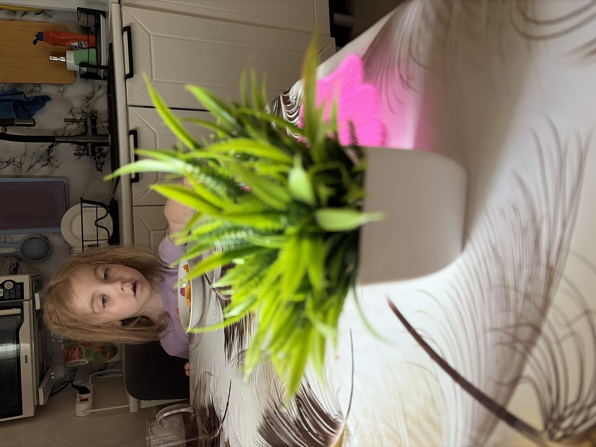 A young girl with light brown hair, her eyes partially closed and mouth open, sits at a table covered with a tablecloth featuring a feather pattern. She is wearing a light purple shirt and is looking down at a white bowl containing food. To her left is a glass pitcher with a dark liquid and a handle. In the foreground, a potted green plant with long, spiky leaves is positioned on the table, obscuring part of the girl and the pitcher. The background shows a kitchen counter with cabinets, a sink, and various kitchen items. The scene appears to be an indoor domestic setting, likely a kitchen or dining area in Tambov, Russia.
