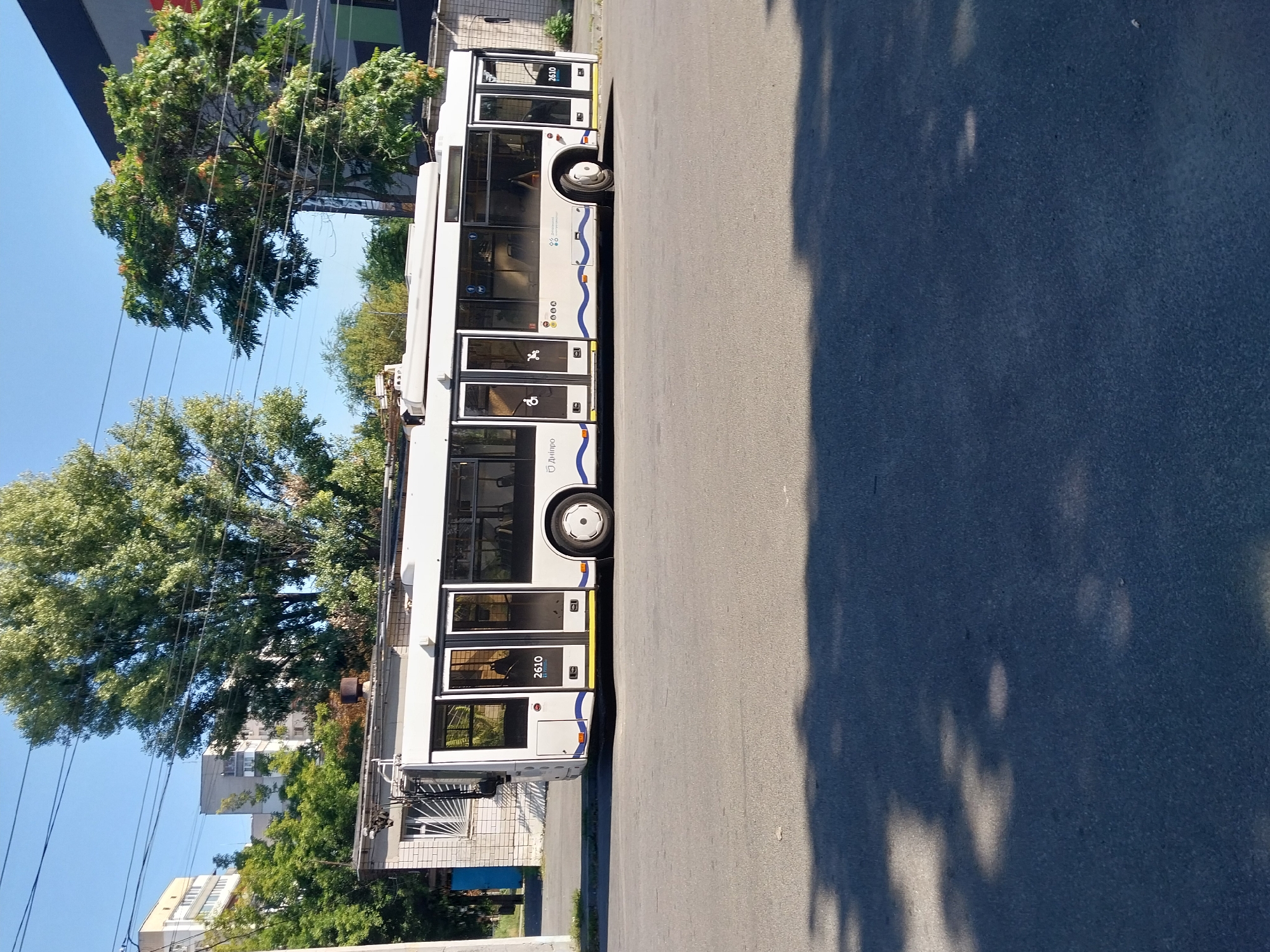 A white trolleybus with the number 2610 is parked on a paved road in the city of Dnipro, Ukraine. The trolleybus is long and modern, with multiple doors and large windows. It is seen from the side, and its front is slightly angled towards the left. The trolleybus displays a blue and white wave-like design along its lower body and the word "ДНІПРО" (Dnipro) in blue lettering on its side.
In the background, there are several buildings, including apartment blocks and what appears to be a low brick structure adjacent to the trolleybus. Trees with green foliage are scattered throughout the scene, with some taller trees framing the sky. Electrical wires are visible crisscrossing above the trolleybus and trees. The ground in front of the trolleybus is asphalt, and shadows from trees are cast across it. The sky is clear and blue, indicating a sunny day. There are no people visible in the image. The scene appears to be a typical urban streetscape.