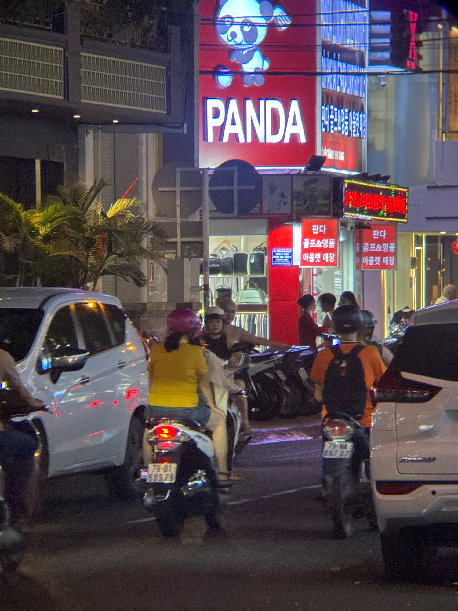 The image depicts a nighttime street scene in Nha Trang, Vietnam, with a focus on traffic and commercial signage. In the foreground, several motorcycles are visible, with riders and passengers. One motorcycle in the center has a license plate reading "79-01 589.79". A white car is parked on the right side of the frame, and a white van is on the left.
Behind the vehicles, a brightly lit storefront features a large red sign with the word "PANDA" in white lettering, accompanied by a cartoon panda image. Smaller red signs with Korean text are also visible on the building, advertising "Golf & Luxury Goods". The street is illuminated by various lights, including neon signs and streetlights, creating a vibrant atmosphere. Palm trees are visible to the left of the storefront. Several people are seen on and around the motorcycles, and some appear to be interacting with each other. The overall environment suggests a busy commercial area within the city.