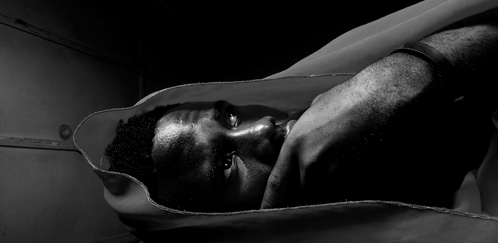 The black and white photograph shows a close-up, angled view of a person's face partially obscured by a draped fabric. The subject, likely a man, has dark skin that is glistening, suggesting sweat or moisture. His eyes are open and looking upwards, with a visible glint of light on his pupils, hinting at a light source positioned above and to his right. The fabric, possibly a hood or garment, is light-colored and loosely covers the upper part of his head and his left arm. A dark band, possibly a bracelet or watch, is visible on his right wrist.
The background is dark and indistinct, with some faint horizontal lines on the left side that might suggest a wall or partition. The lighting is dramatic, with strong contrasts between the illuminated parts of the face and the surrounding darkness, creating a sense of depth and highlighting the textures of the skin and hair. The overall mood appears introspective or perhaps suggestive of a challenging or oppressive environment due to the stark lighting and the way the subject is enveloped by the fabric. The image captures a moment of intense focus or emotion on the subject's face, made more prominent by the high contrast black and white rendering. No text is visible in the image. The location context is Narayi, Nigeria.