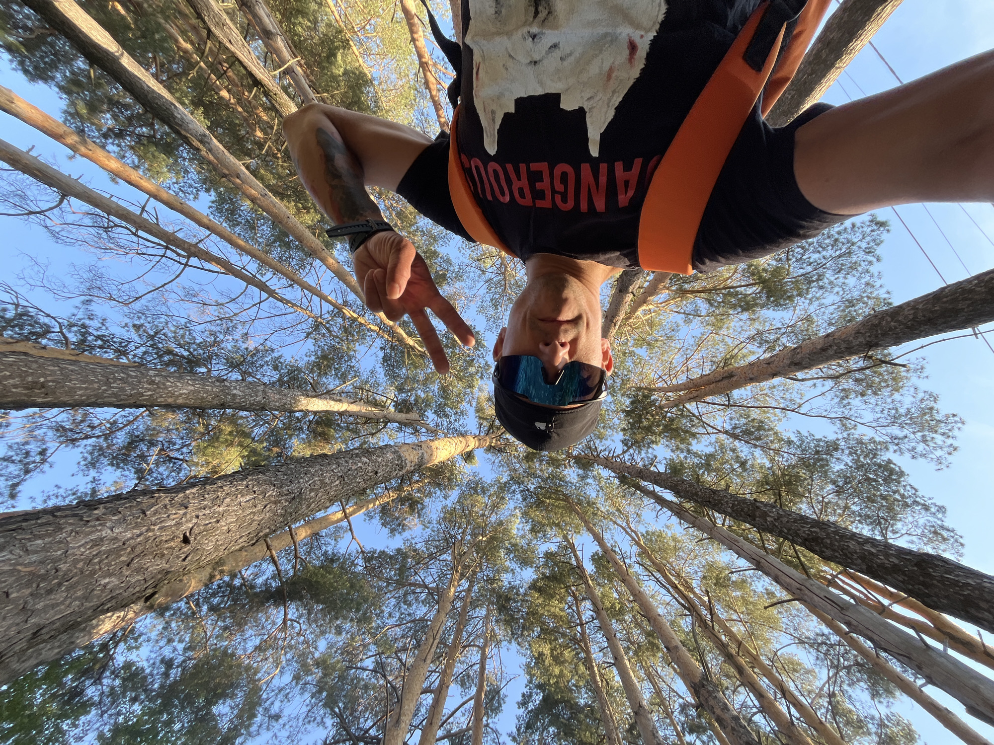 A low-angle shot depicts a man in a black T-shirt with "DANGEROUS" printed in orange, wearing sunglasses and a black cap, giving a peace sign. He appears to be suspended among tall pine trees under a clear blue sky. The location is Chaikovsky, Russia.