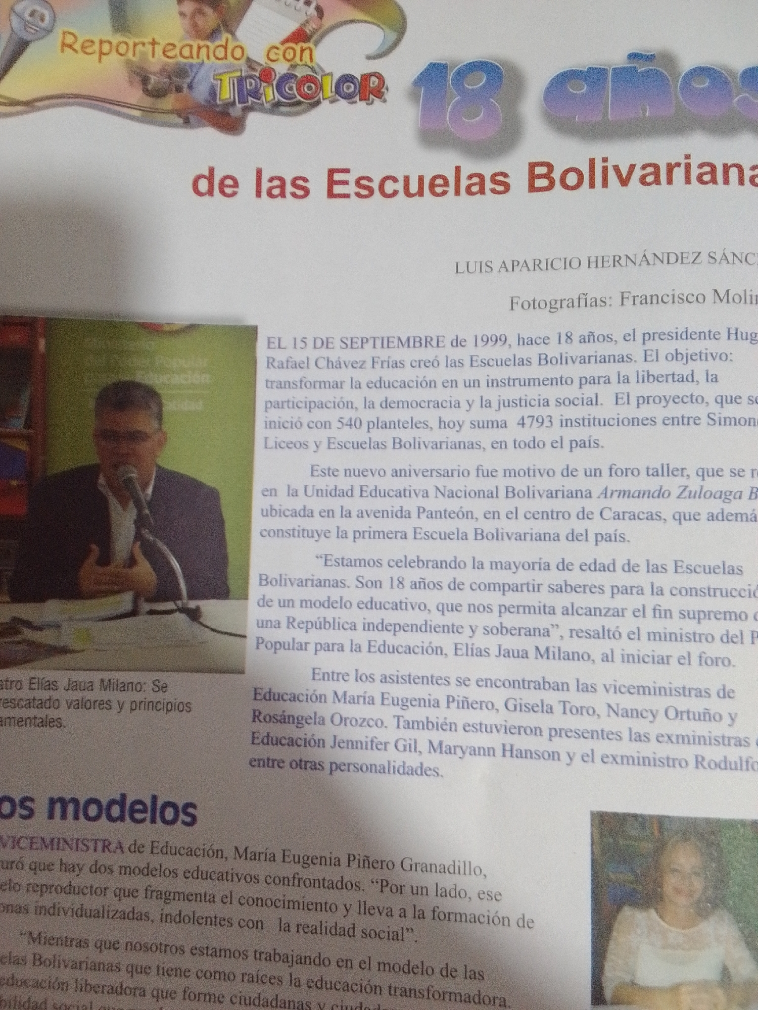 This is a photograph of a newspaper clipping or magazine spread, likely celebrating the 18th anniversary of the Bolivarian Schools in Venezuela.
The primary subjects are two individuals and text. On the left, a man in a dark suit and white shirt is speaking into a microphone, appearing to be at a podium. He is wearing glasses. To the right, a woman in a white lace top is smiling and looking slightly off-camera.
The text prominently features headings and body paragraphs discussing the Bolivarian Schools. Key phrases include "Reporteando con Tricolor," "18 años de las Escuelas Bolivarianas," and names like "LUIS APARICIO HERNÁNDEZ SÁNCHEZ" and "Francisco Molin." The text details the history and objectives of the Bolivarian Schools, mentioning their founding date of September 15, 1999, by President Hugo Rafael Chávez Frías. It also highlights the growth in the number of institutions and quotes a minister discussing the educational model.
The setting appears to be an indoor event, possibly a press conference or forum, given the microphone and podium. The overall tone conveyed by the text is celebratory and informative. The presence of multiple officials, including viceministras and exministras, suggests a significant public event related to education. The image seems to capture a moment of reporting on an anniversary.