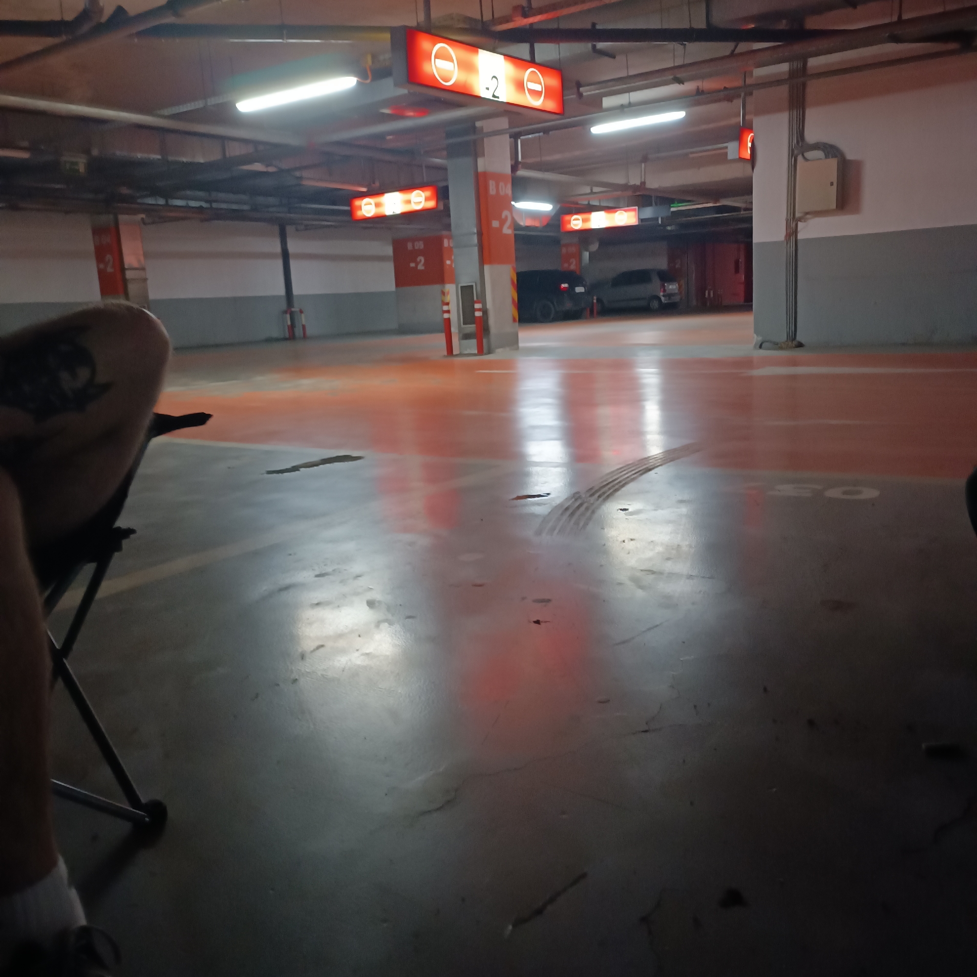 The image captures an underground parking garage, likely located in Sofia, Bulgaria. The primary subject is a person partially visible on the left, sitting in a dark, foldable chair. Their left leg, adorned with a dark tattoo on the thigh, is prominent, along with their lower right leg, wearing a white sock and dark shoe.
The setting is characterized by concrete pillars, an exposed ceiling with various pipes and conduits, and numerous fluorescent light fixtures. The floor is a mix of grey concrete and sections painted in a vibrant orange-red, which strongly reflect the overhead lighting and the glowing red signs.
Several red, illuminated signs are suspended from the ceiling and attached to pillars. These signs feature the international "No Entry" symbol (a red circle with a white horizontal bar) and the number "-2", indicating the parking level. One prominent sign also displays "B 04", suggesting a specific section or block within the garage. Other pillars partially show "B 05" and "B 04" markings.
In the background, a few dark-colored cars are parked, hinting at the garage's function. An electrical box or panel is visible on a grey and white wall section to the right. The floor shows tire marks and some dark stains. The overall lighting is artificial and bright, creating strong reflections on the glossy floor surface. No specific activity or event is clearly depicted beyond the person sitting, possibly waiting or resting. The time of day is not discernible due to the indoor setting.