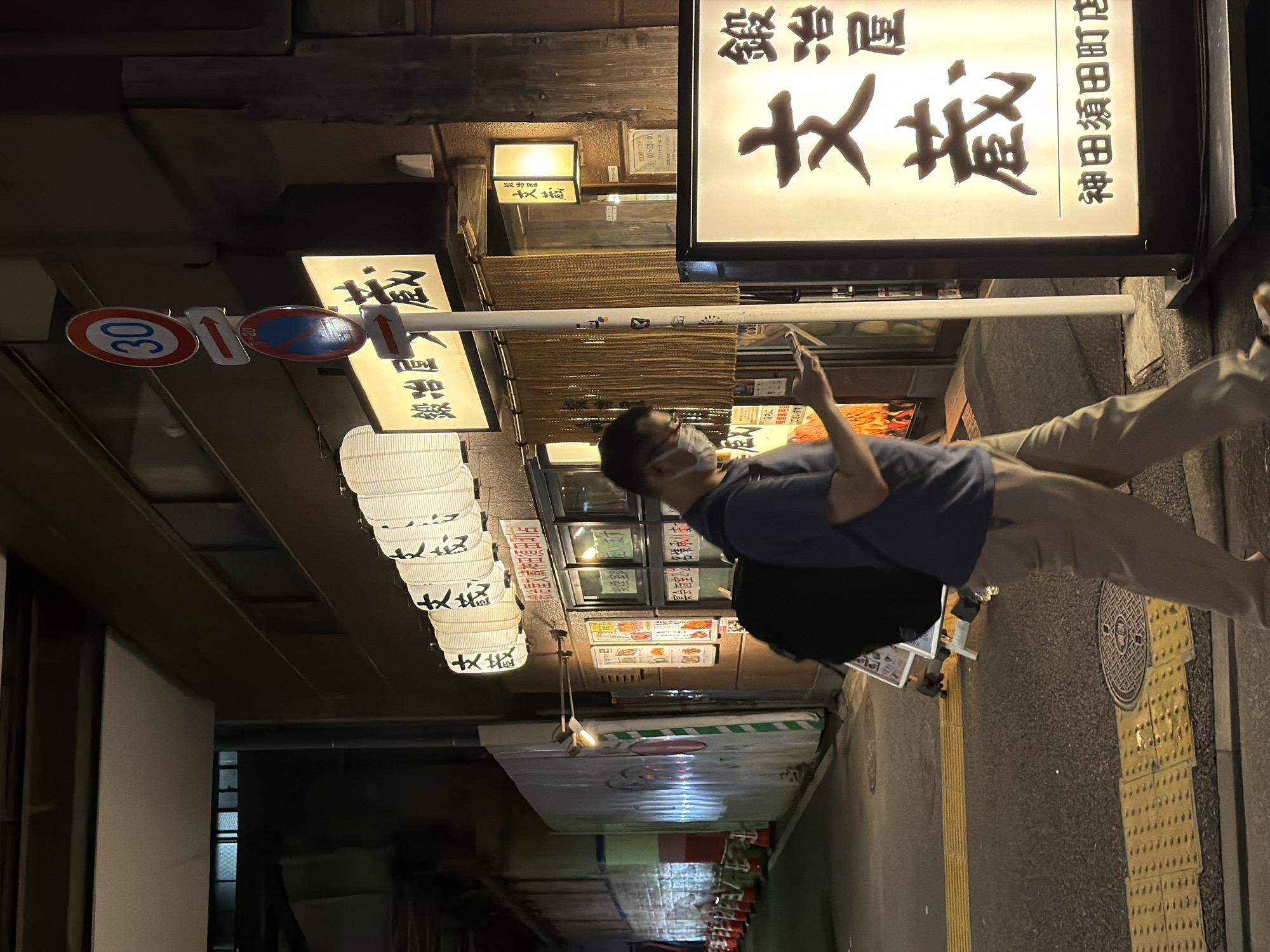 The image depicts a street scene at night in Chiyoda, Japan, likely near a restaurant. A man wearing a blue t-shirt, tan pants, and a face mask is walking while looking at his phone. He carries a black backpack. To his right, a lit sign displays Japanese characters, identifying a business named "文蔵" (Bunzo) with an address "神田須田町店" (Kanda Suda-cho Store). In the background, there are white paper lanterns hanging, another illuminated sign with Japanese text, a beaded curtain, and a menu displayed on the wall. Traffic cones and a construction barrier are visible further down the street. A traffic sign with "30" and an arrow, and another sign prohibiting parking are mounted on a pole. The ground is paved with asphalt and features a tactile paving strip.