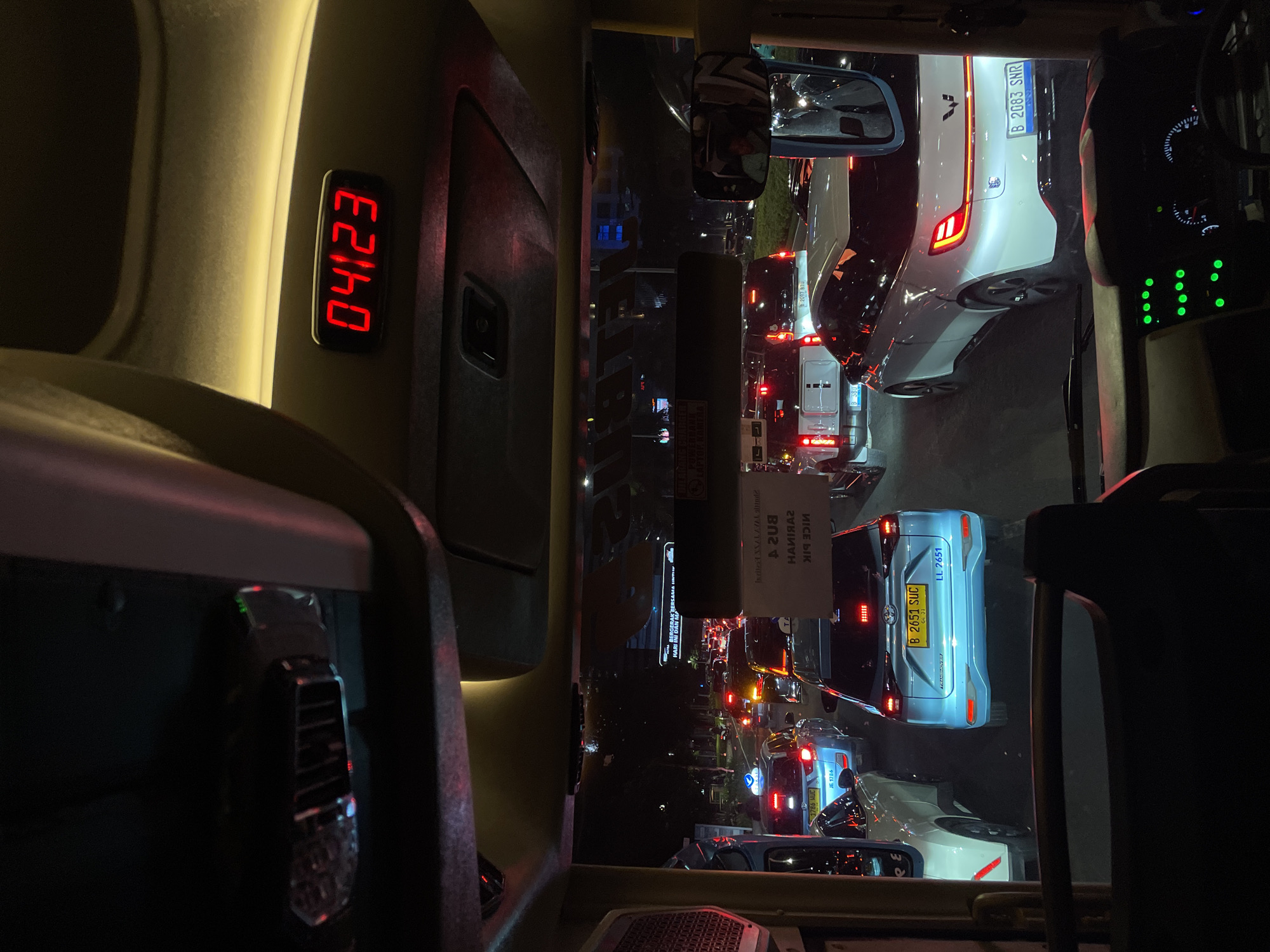 The image, rotated 90 degrees counter-clockwise, shows a nighttime view from the inside of a vehicle stuck in traffic. The digital display shows "04:23". Through the windshield, a dense traffic jam is visible with numerous cars and tail lights. License plates from two cars are visible: "B 2083 SNR" on a white car and "B 2651 SUC" on a light blue car. A sign with some text is present, including "HARI INI BERGERAK BERSAMA". Another sign includes "Bus 24R" and the text "NICE BIK" written backwards. License plate "JE 1786" is also visible. The location appears to be in the city of Central Jakarta, Indonesia, based on the license plate codes "B" and "JE."
