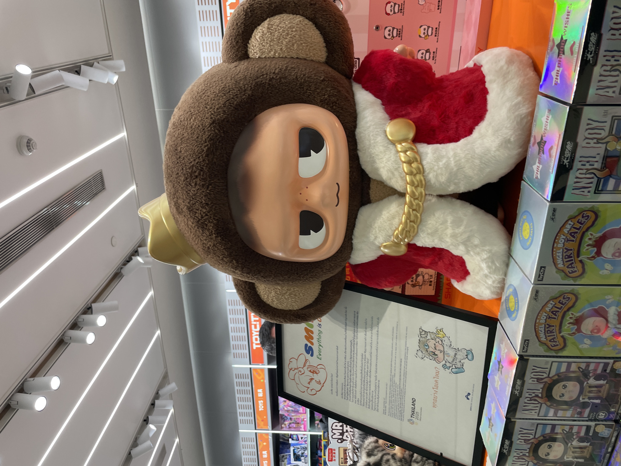 The image depicts a retail display in a shop, featuring a large, plush figure of a character with a bear-like head, a peach-colored face, and a gold crown. The figure wears a red and white robe with a gold rope detail. To the right of the figure, there are several boxes of "ANGEL BOY" and "FAIRY TALES" collectibles. In front of the figure is a framed sign with text and an illustration of a character labeled "THAILAND." The text includes the phrase "Everything is OK." The location is identified on the sign as Thailand. The sign includes the text "THAILAND 2025" and a cartoon illustration of a white creature. The background reveals shelves with toys and the word "TOYCITY" in orange and white. The lighting is provided by ceiling-mounted spotlights.