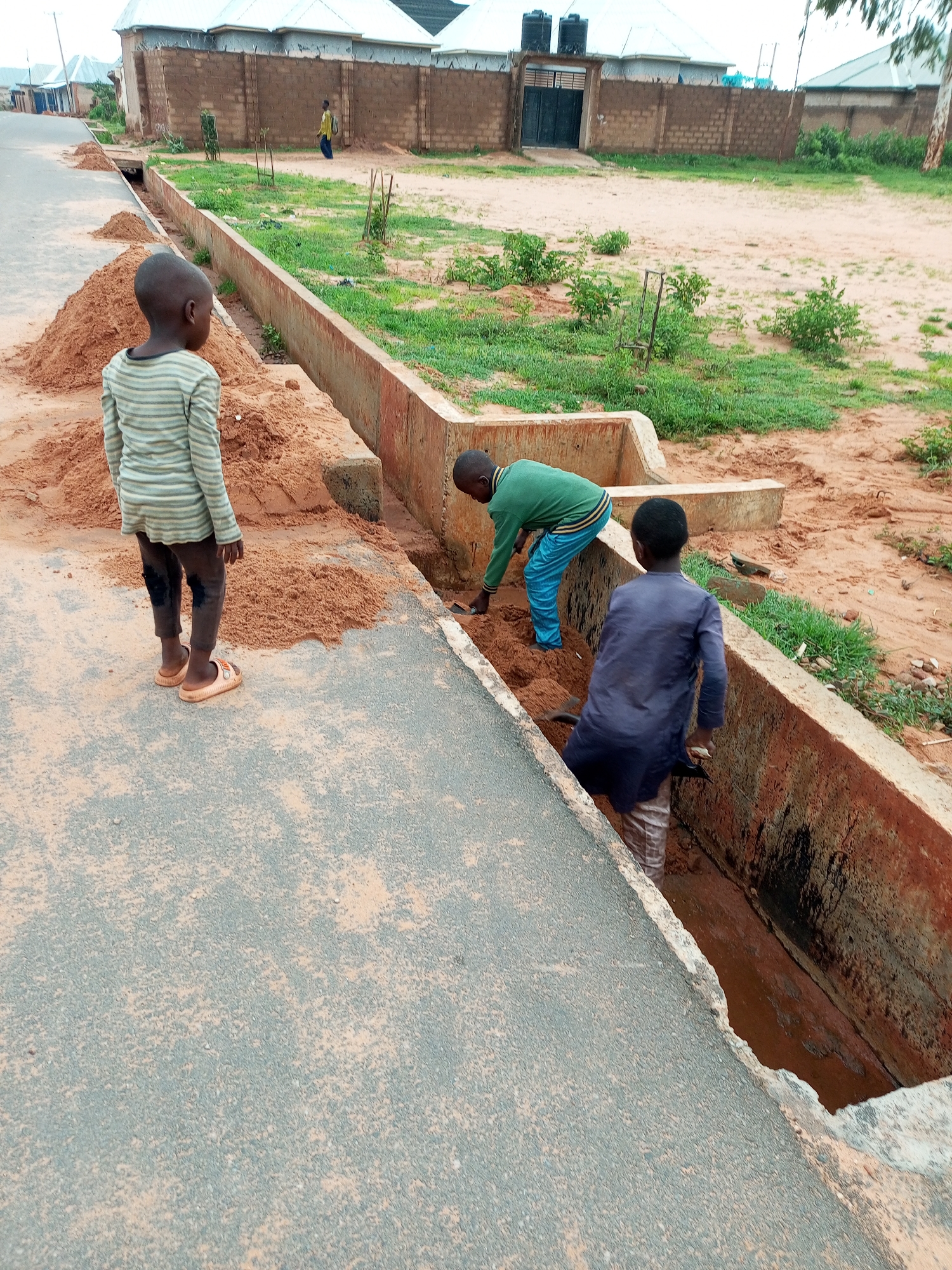 In the city of Gombe, Nigeria, three young boys are engaged in manual labor in an open drainage channel. The scene takes place on a dirt road adjacent to a fenced residential property. The drainage ditch, approximately two feet deep and three feet wide, is filled with reddish-brown soil and some stagnant water at the bottom.
On the left, a boy wearing a striped long-sleeved shirt and dark pants stands on the edge of the road, facing away from the viewer, looking into the ditch. He has light-colored sandals on his feet.
In the middle of the ditch, another boy in a green long-sleeved shirt and blue pants is actively working, digging into the soil with his hands or a small tool. He is bent over, focused on his task.
To the right, a third boy, dressed in a dark blue tunic and striped pants, is also in the ditch. He is partially obscured by the edge of the ditch and appears to be holding a tool, possibly a shovel, as he works on the soil.
Large piles of excavated reddish-brown soil are visible on the side of the road next to the drainage channel, indicating that the boys are clearing it out. The environment suggests a construction or maintenance activity related to the road and drainage system. In the background, a brick wall with a metal gate and two water tanks on top of a building are visible, suggesting the presence of a house or compound. A few small trees and green vegetation are scattered around the area.