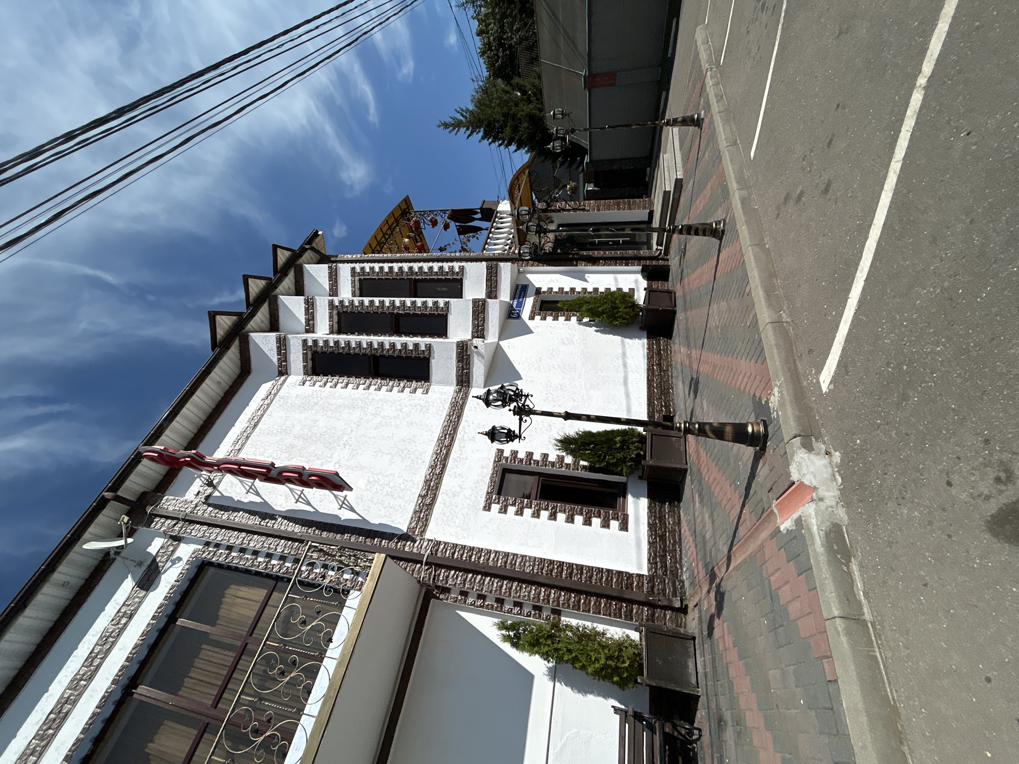 The image shows a commercial building with a white and brown brick facade, a red sign spelling "ОТЕЛЬ" (Hotel) on its upper section, and a balcony with ornate railings on the left side. The building has several dark-framed windows and is flanked by manicured evergreen trees in dark planters. Ornate lampposts are positioned along the sidewalk, which is paved with red and grey interlocking bricks. A street sign with the number "64" and Cyrillic text is visible on the lower part of the building. Beyond the sidewalk, there is a paved area with white parking lines and a grey curb. In the background, a fence, trees, and part of another building are visible under a bright blue sky with scattered white clouds. The scene appears to be an exterior view of a hotel or similar establishment in the city of Novoivanovskoe, Russia, on a sunny day. There are no people visible in the image.