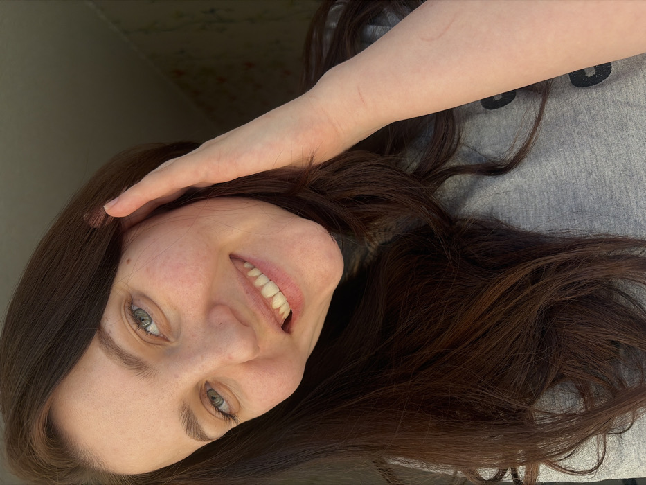 A smiling woman with light skin, brown hair, and gray-blue eyes is angled toward the viewer. Her right hand rests on her forehead. She has a slight smile, revealing straight, white teeth. She appears to be wearing a gray shirt with dark circles printed on it. The background includes a neutral wall with hints of floral wallpaper. The image's orientation suggests it may be a self-portrait or taken from an unusual angle.