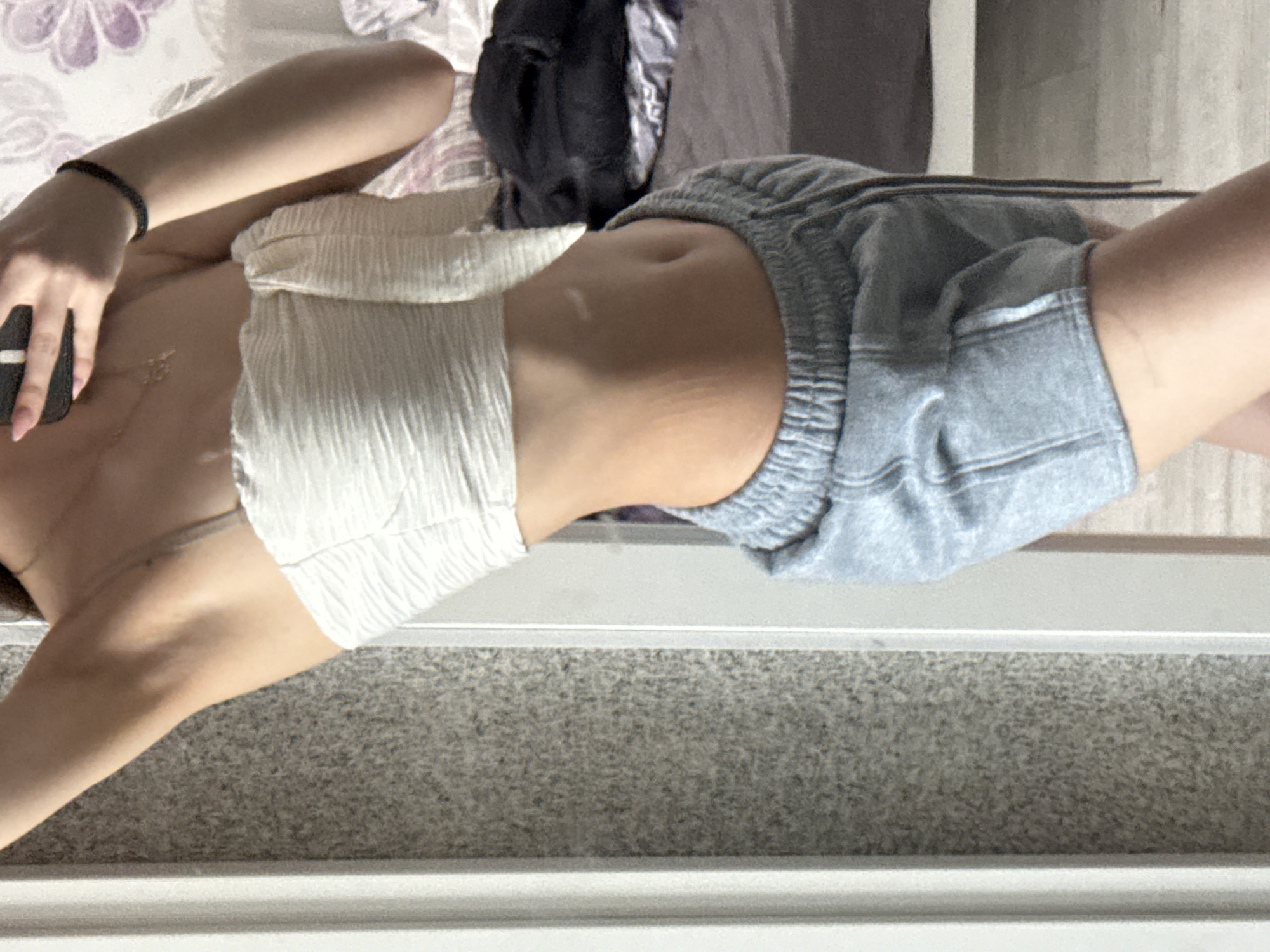 A person with light skin tone is shown in a partial view, wearing a light-colored, textured, strapless top and gray athletic shorts. The person is holding a black object in their hand. The background consists of a bed with floral-patterned bedding, a dark-colored bag, and a gray wall or flooring. The bottom of the image is dominated by a grainy gray surface with a white trim. There is no evidence to suggest that this image was taken in Sofia, Bulgaria.