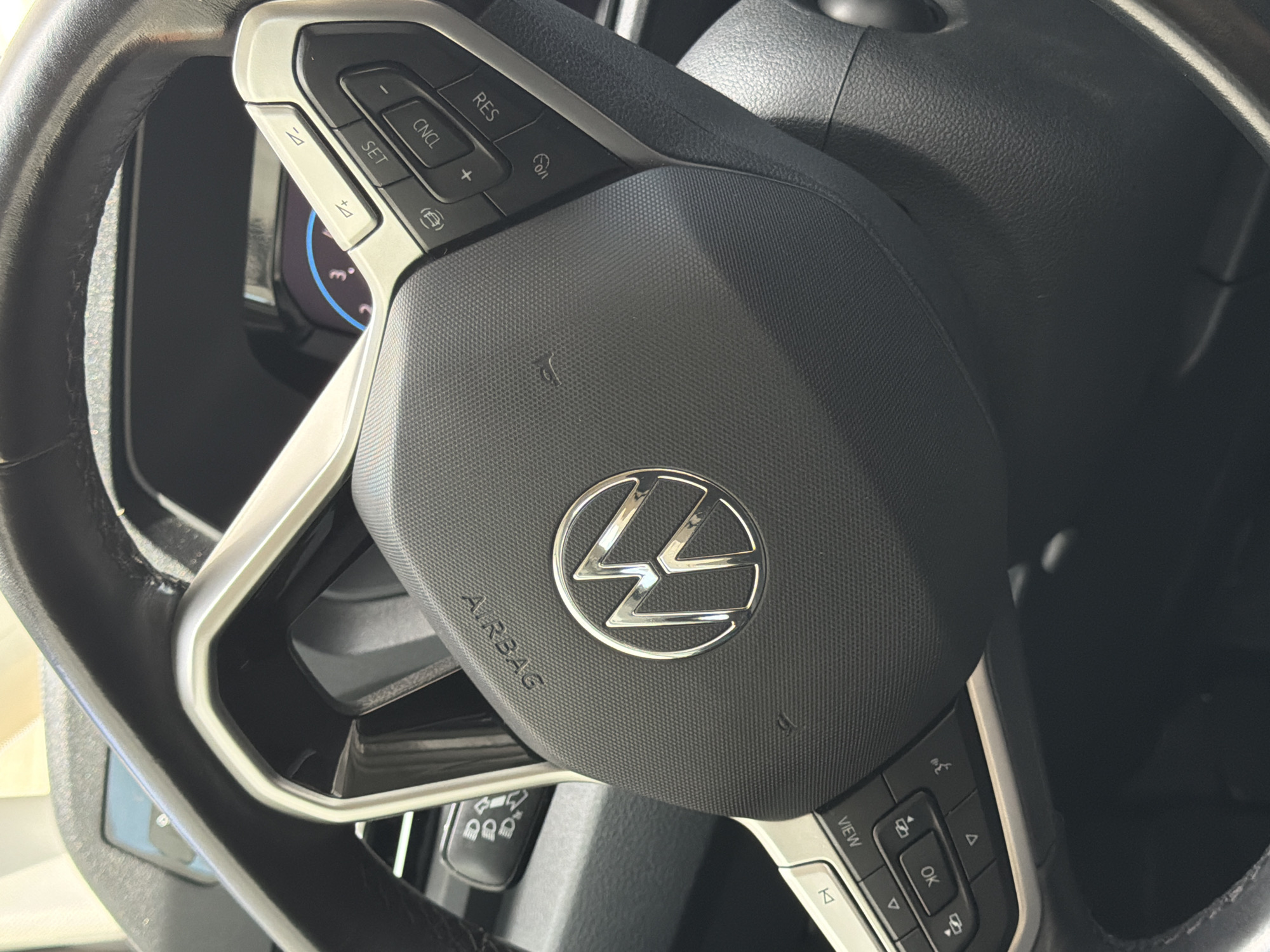 The image shows a close-up view of a car's steering wheel. The steering wheel is black with silver accents and features various control buttons. Prominently displayed in the center of the steering wheel is the Volkswagen logo. To the left of the logo, the word "AIRBAG" is visible.
On the left side of the steering wheel, there are buttons labeled "SET", "CNCL", and "RES". There are also buttons with plus and minus symbols, as well as a button with an "i" symbol. On the right side of the steering wheel, there are buttons labeled "VIEW" and "OK", along with directional arrows.
The background of the image is dark, suggesting the interior of a car. A portion of the car's dashboard is visible on the left side, showing a digital display with some blue and white markings. The image appears to be taken in daylight, as indicated by the lighting. There are no people visible in the image. The setting is likely a parked car.
The visible text includes "AIRBAG", "SET", "CNCL", "RES", "VIEW", and "OK". There are also some symbols on the buttons, including plus, minus, and directional arrows. The Volkswagen logo is also a recognizable symbol. The overall impression is of a modern car's interior, focusing on the driver's controls.
