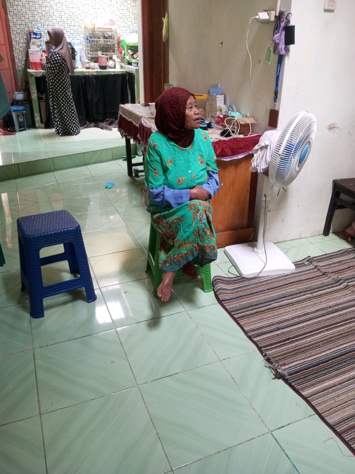 In a tiled room, an elderly woman wearing a red hijab and a green and blue patterned outfit sits on a green plastic stool. She is positioned in the center of the frame, facing to the left. To her left, a blue plastic stool stands empty. Behind her, a white fan is visible, and to her right, a striped rug lies on the floor. The background shows a kitchen counter with various items, and a woman in a black and white polka-dot dress is standing in what appears to be a doorway. The room is lit by artificial light. This image was taken in Krasak, Indonesia.