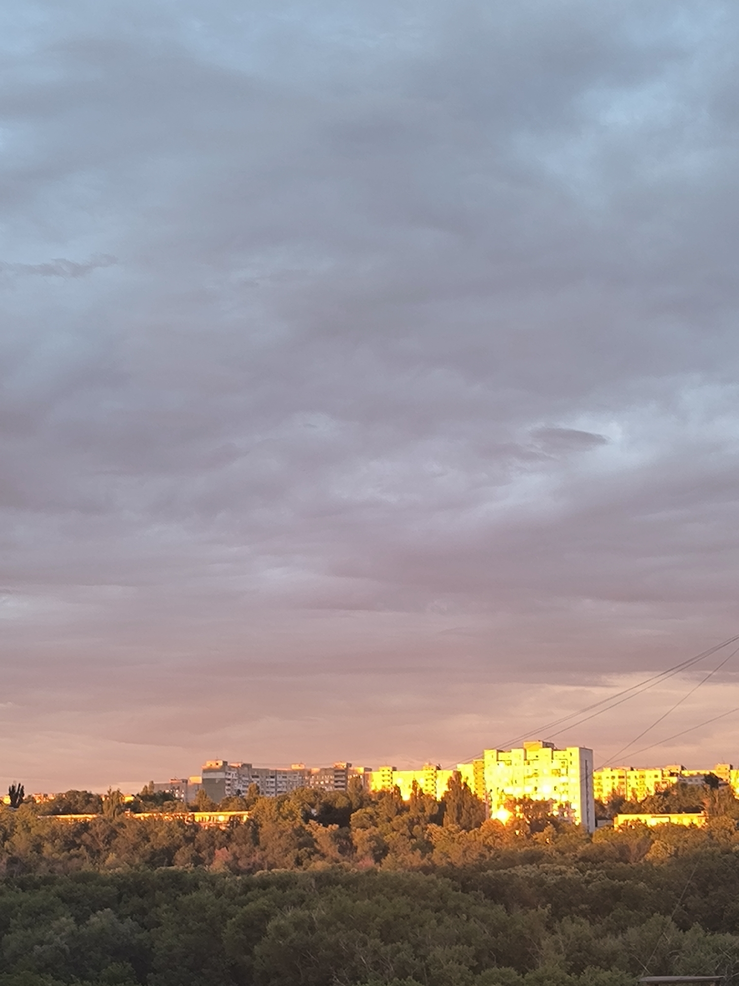 The image shows a cityscape at sunset, with apartment buildings illuminated by the golden light. In the foreground, a dense forest stretches across the bottom of the frame, its dark green foliage contrasting with the brightly lit buildings in the middle ground. The sky above is filled with dramatic clouds, tinged with pink and orange hues from the setting sun. The architecture of the buildings suggests a Soviet-era housing block, typical of many residential areas in Chișinău, Moldova. The scene captures a serene moment as day transitions into night, with the natural beauty of the sunset casting a warm glow over the urban environment.