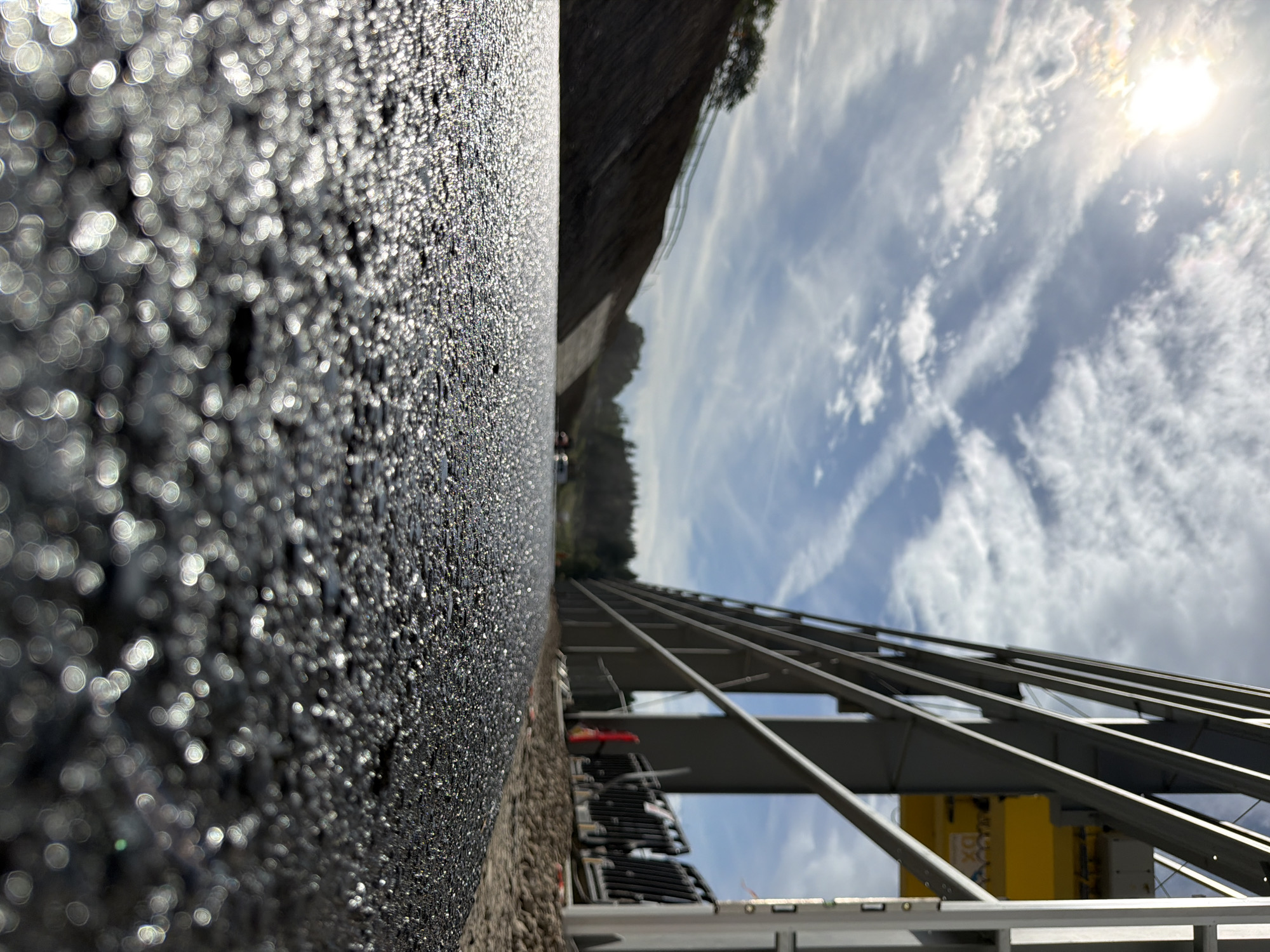 The image shows a low-angle perspective of a newly paved asphalt surface, likely a road or construction site, in Janowice, Poland. The asphalt is glistening with sunlight, creating numerous bokeh effects in the foreground. In the middle ground, a construction site is visible with a large yellow crane and grey metal framing. A sloped embankment covered in dark soil is on the left, with trees and foliage in the distance. The sky is blue with scattered white clouds and the sun is bright and partially obscured by clouds. The scene appears to be a construction project in progress.