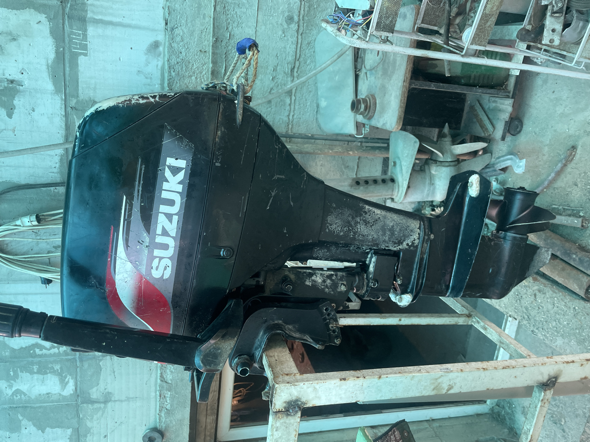 The image shows a black Suzuki outboard motor resting on a white metal frame, likely a stand. The motor displays some wear and tear. In the background, there are various metallic objects, including what appears to be a propeller. The environment includes a concrete wall. There is no indication of people or actions. There is no way to verify or ascertain the location as Olginka, Russia, based on the image alone.