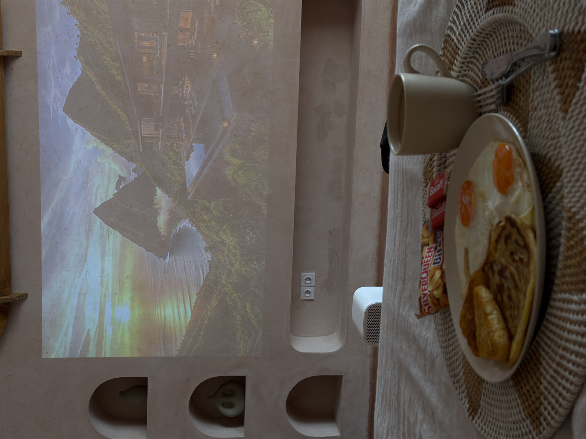 A meal of eggs and pastries with a cup of coffee and candy rests on a circular woven placemat on a table. To the left, a nature scene appears on a wall, possibly a projection. The wall has built-in alcoves and a light switch. The scene is reminiscent of a room, but there is no evidence to confirm its location in North Kuta, Indonesia.