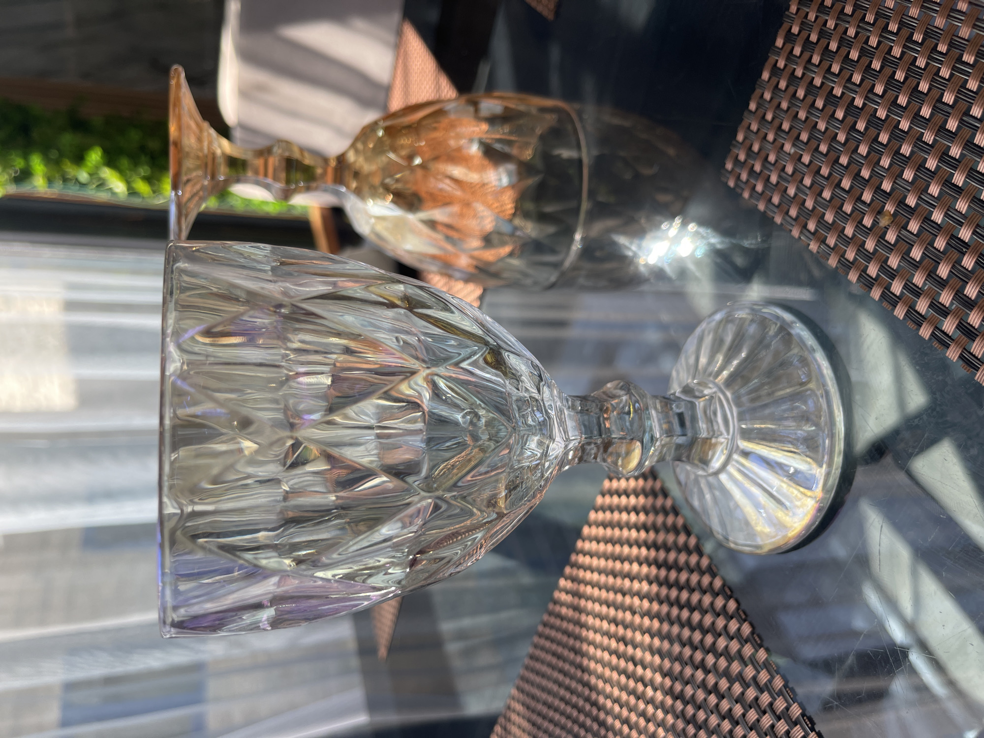 The image shows two stemmed glasses on a dark, reflective table. The glass in the foreground is clear and has a diamond-cut pattern. It is standing upright. The glass in the background is amber-colored and is inverted, resting on its rim. Both glasses are placed on a woven placemat with a brown and tan pattern. In the background, sheer white curtains are visible, and beyond that, blurred greenery and sunlight suggest an outdoor setting. The scene is lit by natural light, casting shadows and reflections on the table surface. The location is specified as Bosteri, Kyrgyzstan.