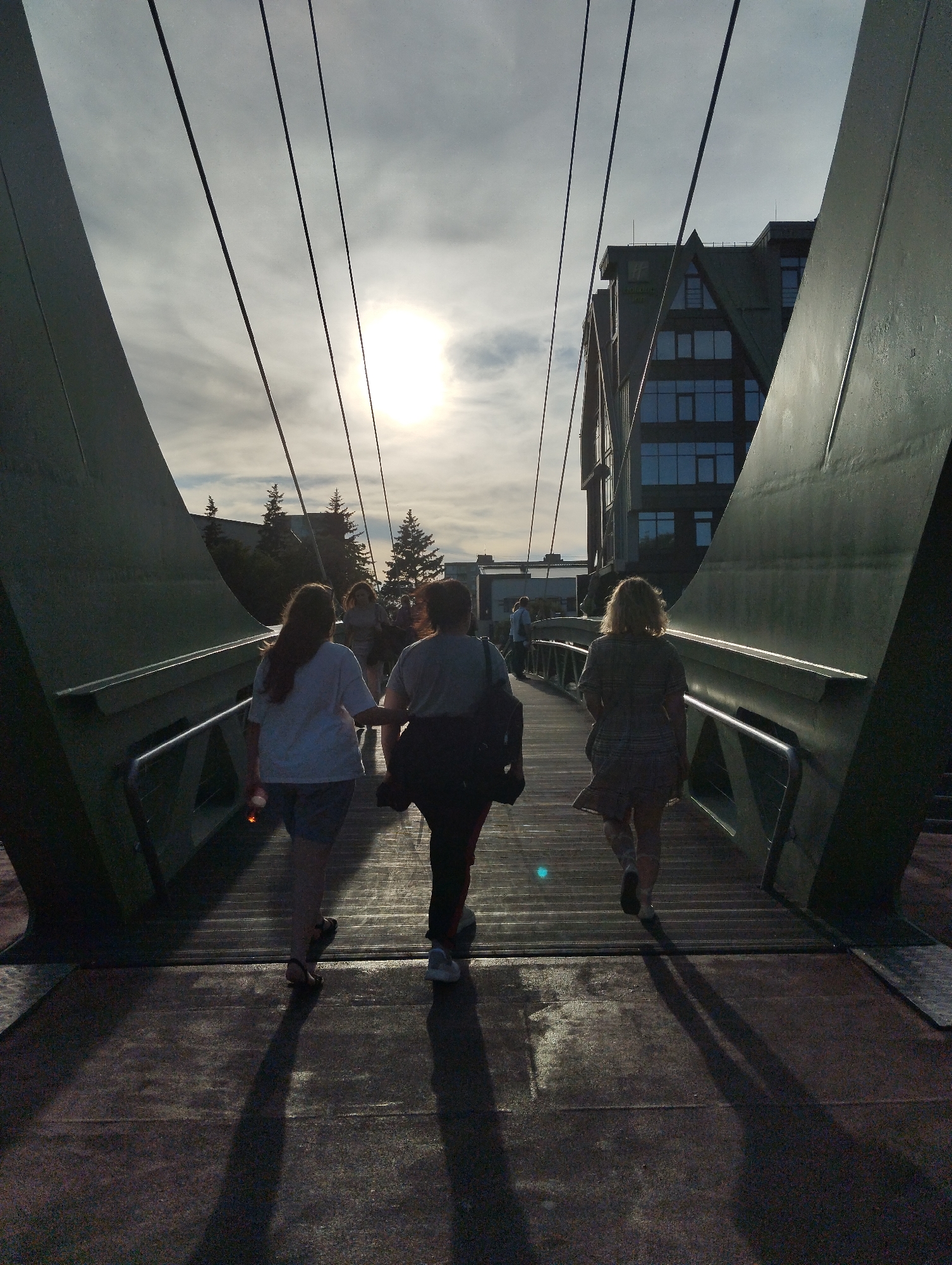 This is a low-angle, natural light photograph of three people walking away from the camera on a bridge. The sun is visible in the upper center of the frame, suggesting it is either sunrise or sunset. The sky is partly cloudy. The bridge appears to be modern, with large green structural elements on either side and thin cables overhead. The walkway is made of wooden planks.
In the foreground, on the left, a woman with long, dark hair is wearing a white t-shirt and dark shorts. She is holding a bottle in her right hand. To her right, a person wearing a gray t-shirt and dark pants is walking. Further ahead, on the right, a woman with blonde hair is wearing a plaid dress and carrying a bag over her left shoulder. The three people are walking in the same direction.
In the background, trees and a multi-story building are visible. The building has a distinctive, angular design with multiple windows. The overall mood of the image is serene, with the warm sunlight casting long shadows on the bridge. The location is identified as Kaliningrad, Russia. There is no visible text in the image.