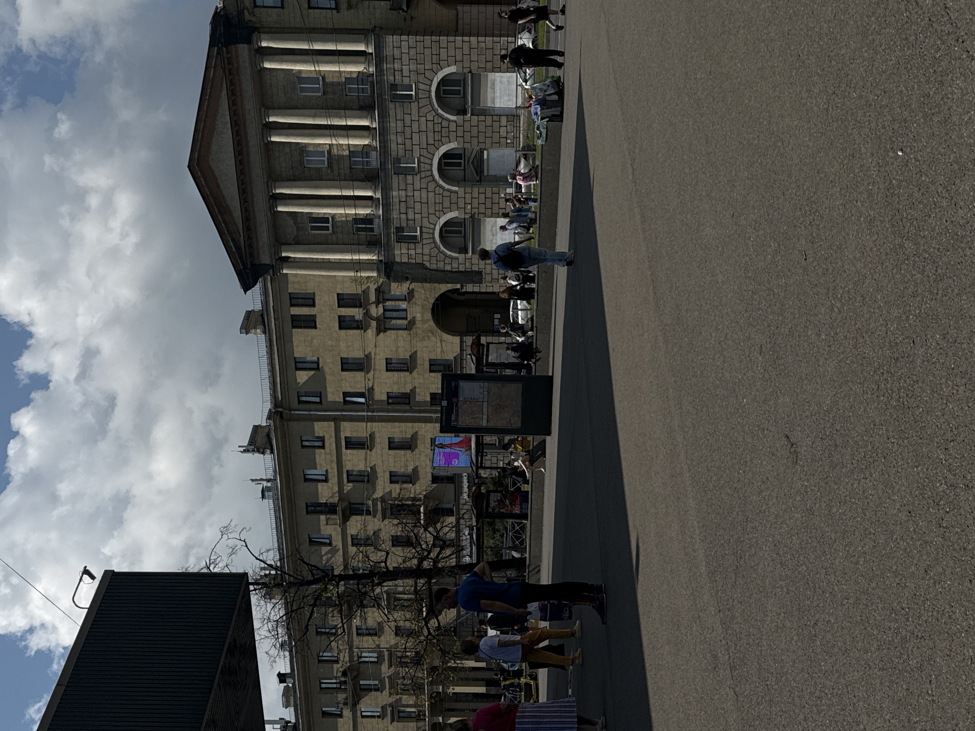 The image captures a street scene in Saint Petersburg, Russia, likely in the vicinity of Nevsky Prospekt, given the architectural style. In the foreground, a wide asphalt area, possibly a pedestrian zone or plaza, dominates the lower half of the frame. To the left, a large, dark, modern rectangular structure juts into the frame from the edge. Behind this, a multi-story building with a light-colored facade and numerous windows stretches across the middle ground. Adjacent to this, a grander, classical-style building features a portico with large columns and a triangular pediment. Scattered across the scene are several individuals, some walking, others standing or sitting. There appears to be a small vendor stall with clothing or textiles displayed on a cart. The sky above is partly cloudy with patches of blue. The overall impression is of a public urban space on a bright day.