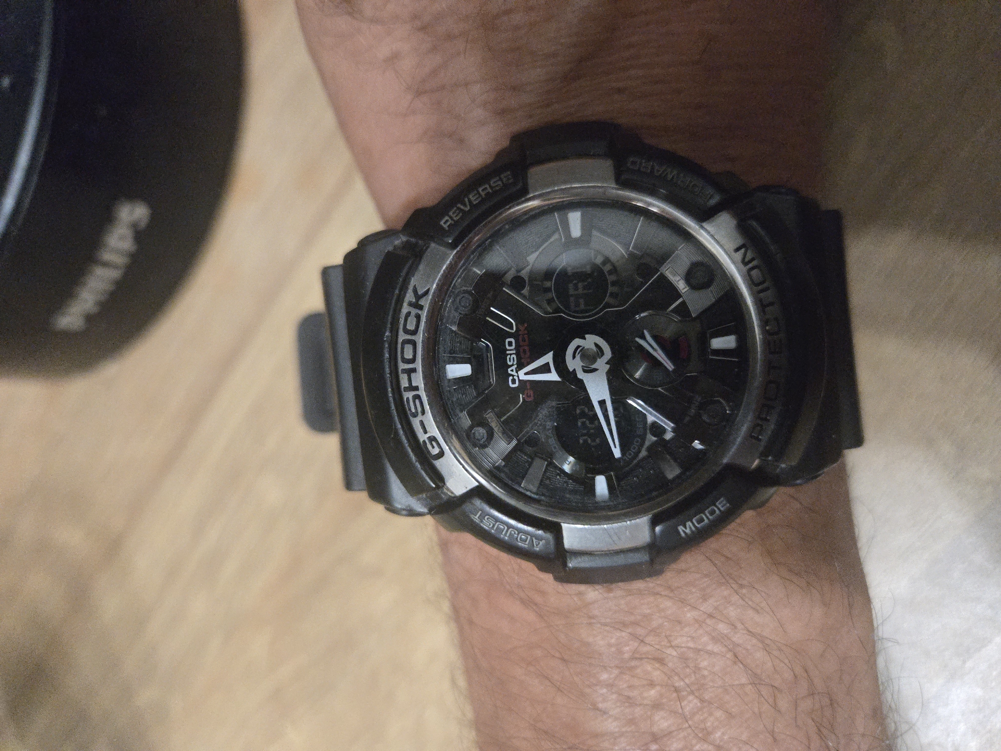 A close-up shot shows a Casio G-Shock watch strapped to a hairy arm. The watch face is black with white hands, displaying the time. The G-Shock logo is prominent on the side of the watch. To the left of the watch, partially out of focus, is a Philips brand product. The background is a light-colored wooden surface. There is no information about the location of the city of Unknown, Unknown.