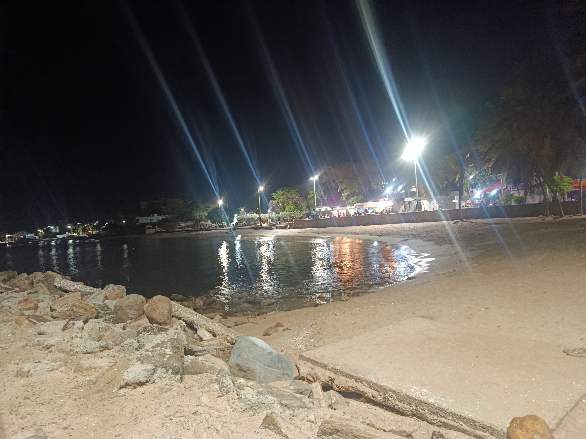 This image captures a nighttime coastal scene in Juan Griego, Venezuela, looking across a calm bay towards an illuminated shoreline. In the immediate foreground, a rocky embankment consisting of large, irregular stones and concrete debris occupies the left side, transitioning into a sandy beach area on the right, which is bordered by a concrete edge.
The water of the bay, dark and exhibiting subtle ripples, reflects numerous artificial light sources from the opposite shore, creating elongated, shimmering patterns. Along the middle ground of the shoreline, multiple bright streetlights emit intense white light, generating prominent vertical lens flares that streak upwards and across the frame. These lights illuminate a promenade or street lined with trees, including palm trees, which obscure portions of the structures behind them.
Under the bright illumination, several indistinct human figures are visible, some appearing seated at outdoor establishments or walking, indicating social activity. These establishments display various internal lights, contributing warm orange and yellow reflections on the water. In the background, further along the distant shore, additional buildings and scattered lights are discernible against the dark night sky. The sky itself is uniformly dark, offering no visible celestial details.