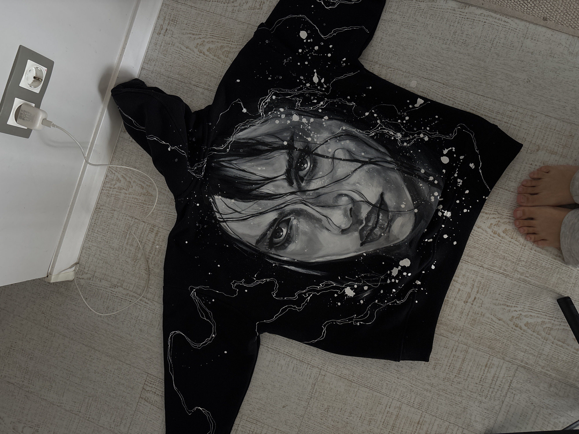 The image depicts a black hoodie with a grayscale portrait of a woman painted on the front. The hoodie is lying on a light-colored wooden floor, next to a white wall. A wall outlet with a charging cable plugged into it is visible on the left. A pair of bare feet belonging to a person wearing gray sweatpants is visible in the top-right corner of the frame. A black object, possibly part of a camera tripod, is visible near the feet. The location is indoors. There is no evidence to suggest the location is in the city of Krasnoyarsk, Russia.