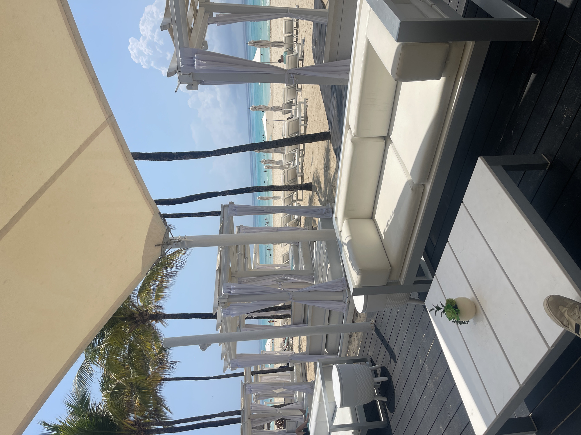 The scene is a high-end beach club in Playa del Carmen, Mexico. The perspective is from under a large beige sun shade. A row of white cabanas with draped white curtains leads toward the beach and ocean. Beyond the cabanas are numerous white lounge chairs and umbrellas on the sand. The turquoise ocean is visible in the distance, with some people in the water. Closer to the viewer, there is white outdoor furniture including a large sofa, a coffee table with a potted succulent, and a small round side table. The furniture sits on a black wooden deck. A portion of a foot and shoe are visible in the bottom right corner of the image. Palm trees are scattered throughout the scene. The sky is blue with scattered clouds.