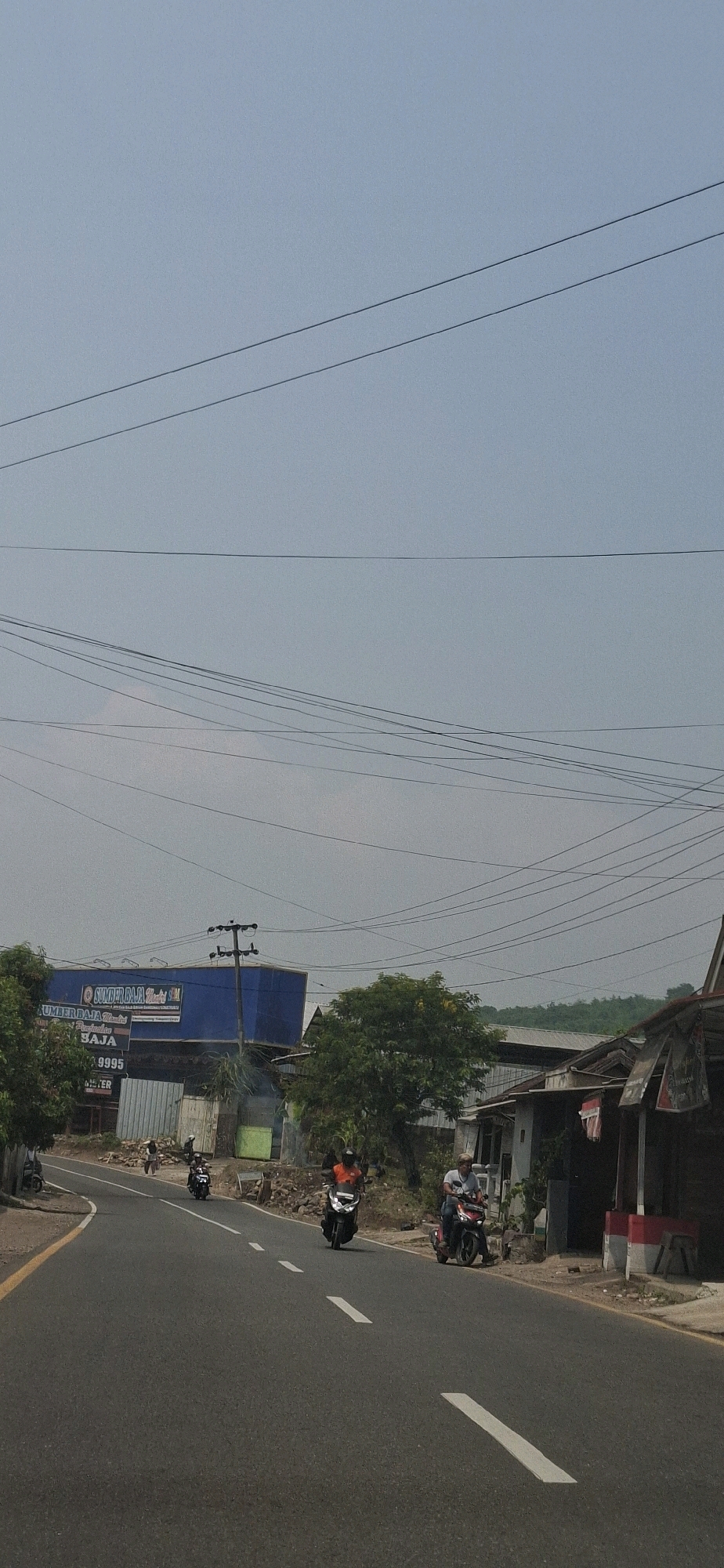 The image shows a street scene in Susukan, Indonesia, with several motorcycles on the road. On the left side of the road, there is a blue building with signage that appears to be a store or business. Trees and smaller buildings are visible along the right side of the road. Overhead, a complex network of power lines is strung across the sky. The sky is overcast, and the overall lighting suggests it is daytime. Several people are riding on the motorcycles, appearing to travel along the road. The road itself is paved and marked with white lines.