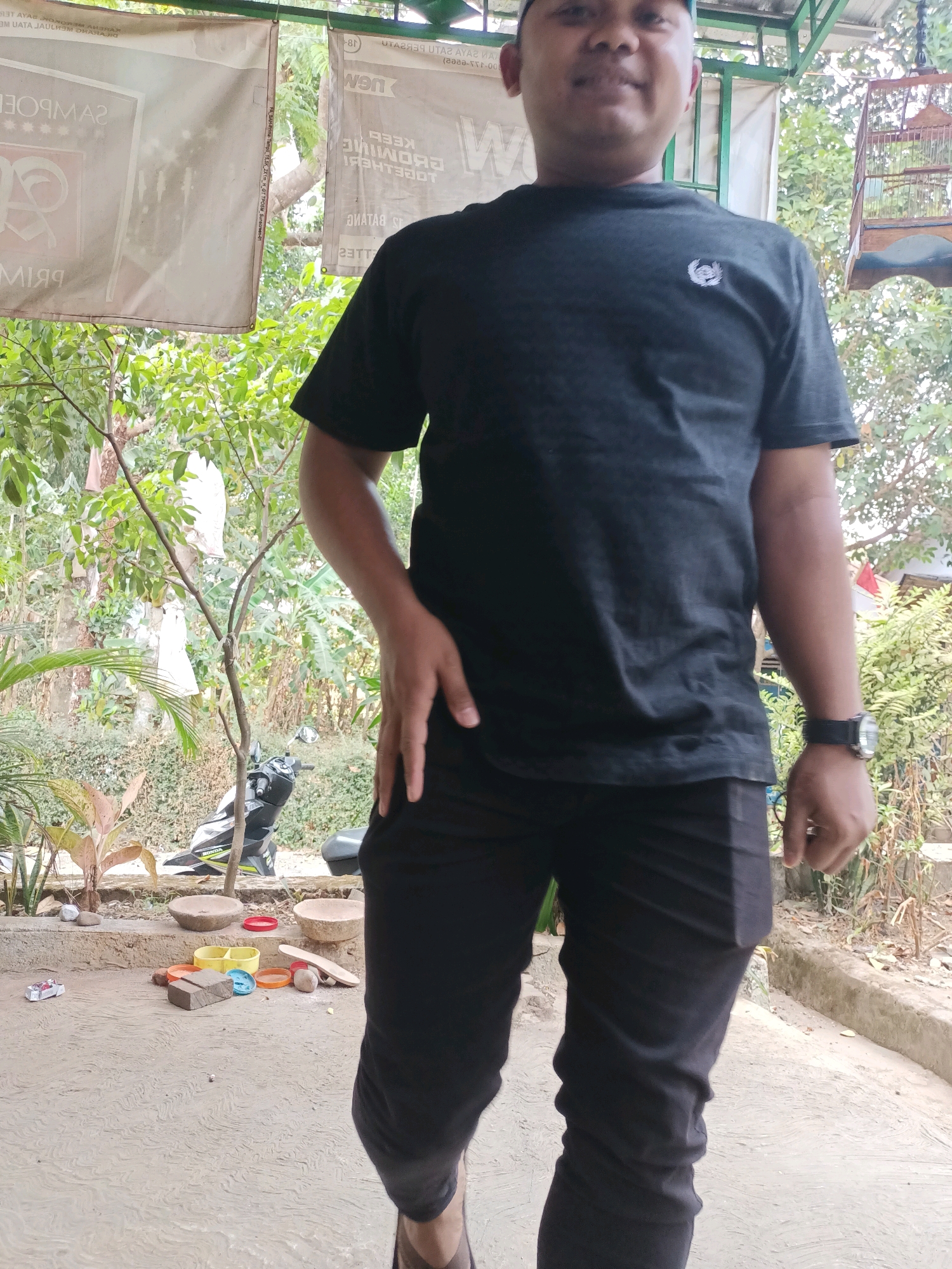 A full shot shows a man standing in what appears to be an outdoor covered area, possibly a carport or open-air building, in Warungpring, Indonesia. He is wearing a black t-shirt with a small white emblem on the left breast and black pants. His right hand is resting on his hip, and he is wearing a watch on his left wrist. The man is facing forward with a neutral expression.
The environment includes a concrete floor with a textured, wavy pattern. Behind the man, the background consists of lush green foliage, including trees and bushes. Hanging from a branch of a tree are several white bags or coverings. In the background, there is a birdcage suspended from the ceiling structure, and a portion of a motorcycle is visible on the left side of the image. Scattered on the ground in front of the man are various objects, including a toy car, several colorful plastic items, what appear to be stones or bricks, and a single sandal. Two circular stone bowls are also present on the ground. The overhead structure is made of metal poles and appears to be covered by a green awning or roofing material. Signs with text are partially visible hanging in the background. The lighting suggests it is daytime.