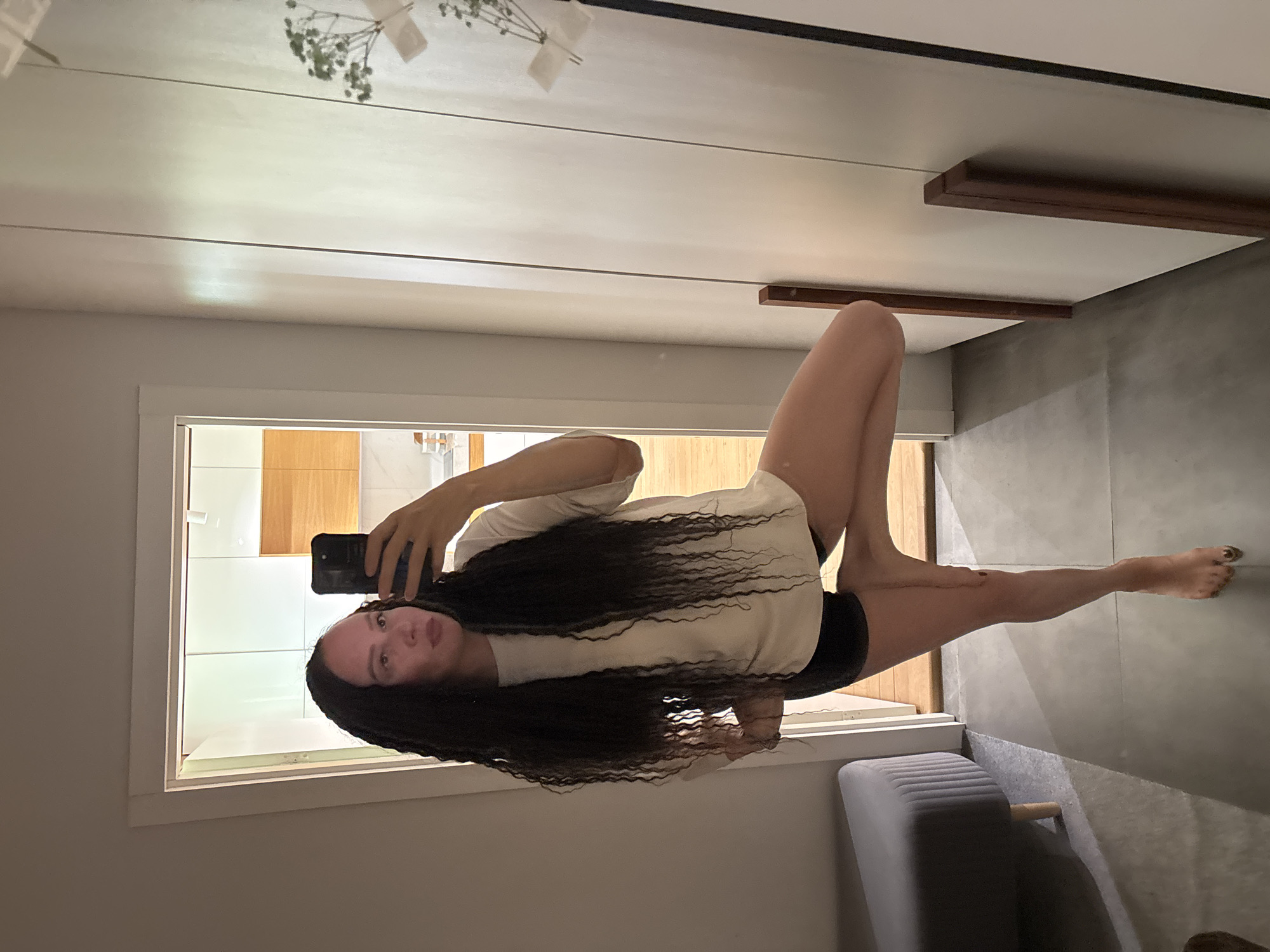 A female subject is captured in a full-body mirror selfie, standing in a yoga pose within what appears to be a residential interior in Chaikovsky, Russia. The subject has long, dark, wavy hair and is wearing a light-colored, short-sleeved t-shirt and dark shorts. Her right foot is placed on her left inner thigh, consistent with Vrksasana (Tree Pose). She holds a black smartphone in her right hand, angled towards the mirror, while her left hand rests on her hip. A faint light pink mark or reflection is visible on her forehead. The subject's reflection occupies the left half of the image.
The environment includes a large, light-colored framed mirror on the right, featuring two dark brown vertical handles or trim pieces. Reflected in the mirror's upper right corner is a small, dried floral arrangement or plant, secured with white tape. To the left of the subject, a gray upholstered bench or ottoman with light wooden legs is partially visible. The floor in the immediate foreground is dark gray, while the floor visible through an open doorway behind the subject is light wood. The doorway opens into a room with light wooden cabinetry and white upper cabinets or walls, suggestive of a kitchen or dining area. A light fixture is visible on the ceiling of the reflected room. The wall on the immediate left (not reflected) is a plain, light color.