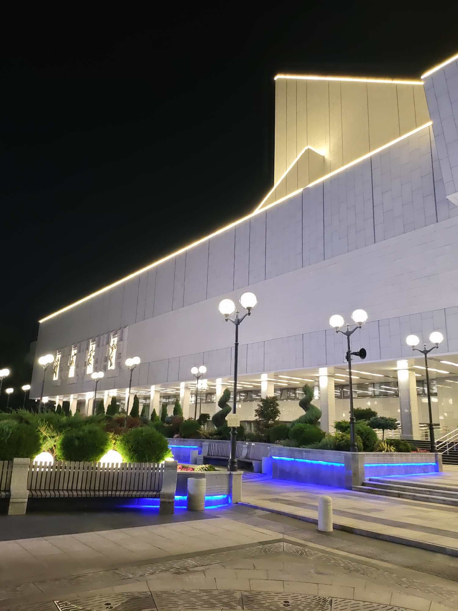 This image depicts the Rostov State Opera and Ballet Theatre, located in Rostov-on-Don, Russia, at night. The building features a modern architectural style with white facade panels and illuminated yellow lights accentuating its angular roofline. In front of the building, there is an outdoor area with paved walkways, benches, and meticulously landscaped bushes and trees. Several streetlights with multiple globes illuminate the scene. The lower sections of the planter boxes and steps are lit with vibrant blue LED lights. The sky is dark, indicating nighttime. There are no people visible in the image.