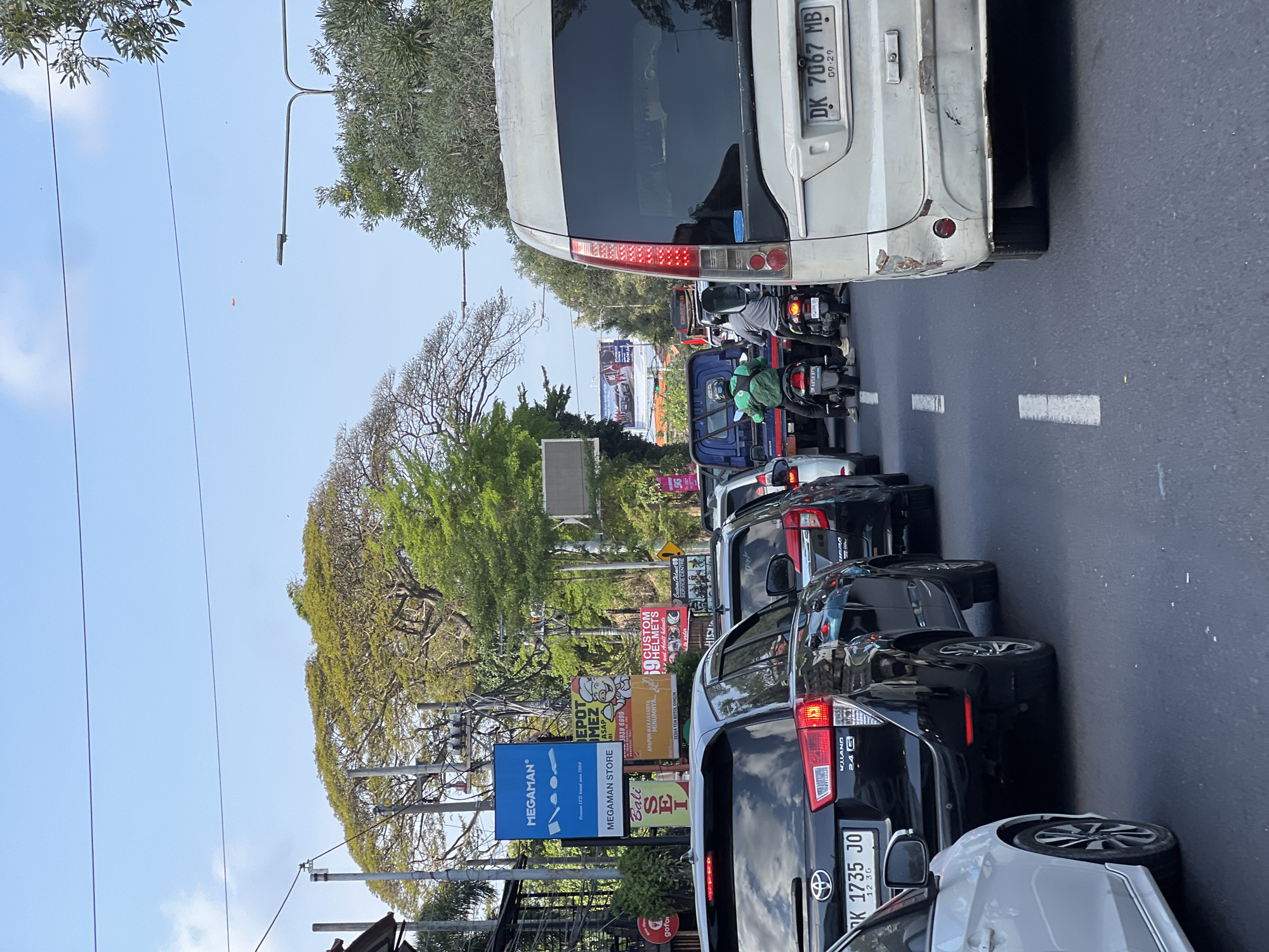 The image depicts a traffic jam on a street in Denpasar, Indonesia. Several vehicles, including cars, motorcycles, and a van, are stopped on the road. A person wearing a green jacket is on a motorcycle in the foreground. Billboards and advertisements are visible on the side of the road, and large trees line the street. The sky is clear and blue. The scene appears to be a typical urban environment during daylight hours. The license plates on the vehicles suggest they are registered in Bali, Indonesia.