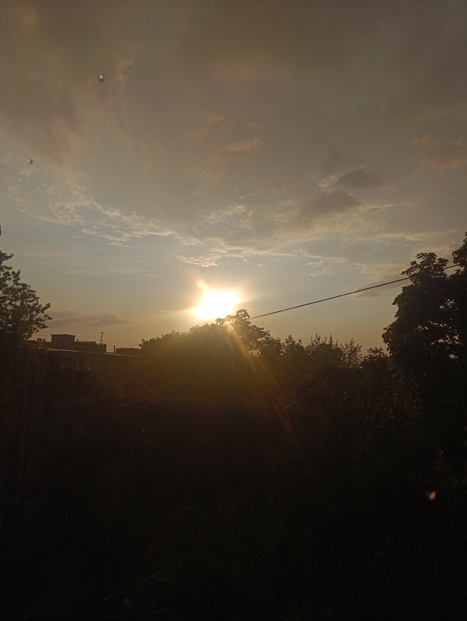 The image captures a sunset scene, likely from the city of Zlatopil, Ukraine, with the sun partially obscured by trees and a building. The sky is filled with a mix of clouds, with a bright solar glare dominating the right side of the frame. A thin wire stretches horizontally across the mid-section of the image, possibly a power line. In the lower portion, a silhouetted building with visible windows and rooftop structures is visible, along with dark foliage and trees. The overall lighting suggests either early morning or late evening, with the sun's rays casting a warm, golden hue. There are no people or animals visibly present in the scene. The presence of a building suggests an urban or semi-urban environment.
