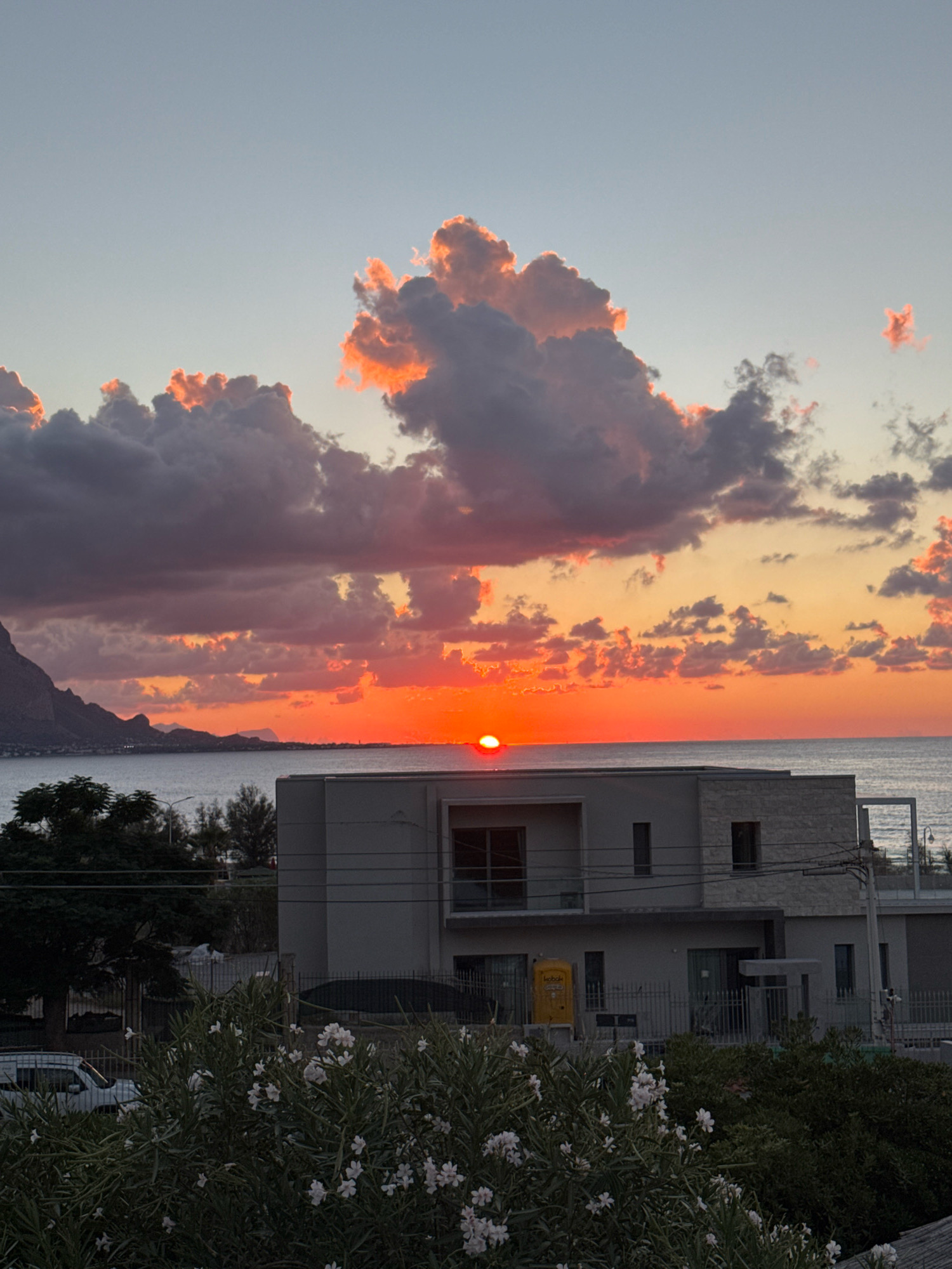 The image captures a sunset over the sea in Capaci, Italy. The sun, a bright orange orb, is low on the horizon, casting a warm glow across the sky and illuminating the undersides of scattered clouds with vibrant hues of orange and pink. In the foreground, a modern building with clean lines and a light-colored facade stands out. Its windows are dark, reflecting the fading light. Surrounding the building, there are trees and lush green foliage, with a cluster of white flowers in the lower right corner. The sea stretches out to the horizon, calm and reflecting the colors of the sky. In the distance, a mountainous coastline is visible, hinting at the unique topography of the region. A white car is parked on the left side of the frame, partially obscured by the greenery. A portable toilet, yellow in color, is positioned near the building. The overall scene suggests a peaceful evening in a coastal Italian town.