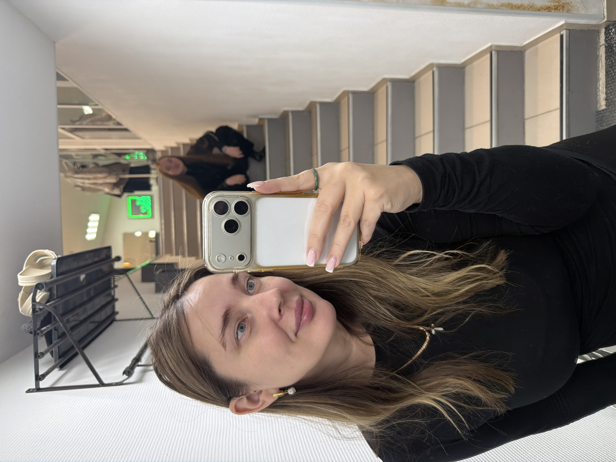 The image depicts an interior scene within a commercial or public building in Chaikovsky, Russia, captured as a mirror selfie. In the foreground, a woman with long, wavy brown hair, blue eyes, and fair skin is visible from the chest up, looking towards the camera with a slight smile. She wears a black long-sleeved top, a gold chain necklace with a small cross pendant, and a gold earring with a white pearl on her left ear; a small stud piercing is visible on her left upper ear cartilage. Her right hand, adorned with a thin green ring on the ring finger and nails featuring a French manicure, holds a light-colored smartphone with a multi-lens camera array, positioned for the selfie.
The background, reflected in the mirror, shows a modern interior space. A staircase with light-colored tiled treads, grey risers, and metallic edge strips ascends to the upper right. On the upper portion of these stairs, a second individual, a young woman with long brown hair and dark clothing, is crouched, looking towards the camera. To the left, a black metal wall-mounted coat rack or shelf holds light-colored items on a textured white wall. Further into the reflection, bright neon green signs, one resembling a hand or arrow, and other overhead lighting fixtures are visible on a distant wall, along with reflections of glass partitions and hanging garments, suggesting a retail or service-oriented environment.