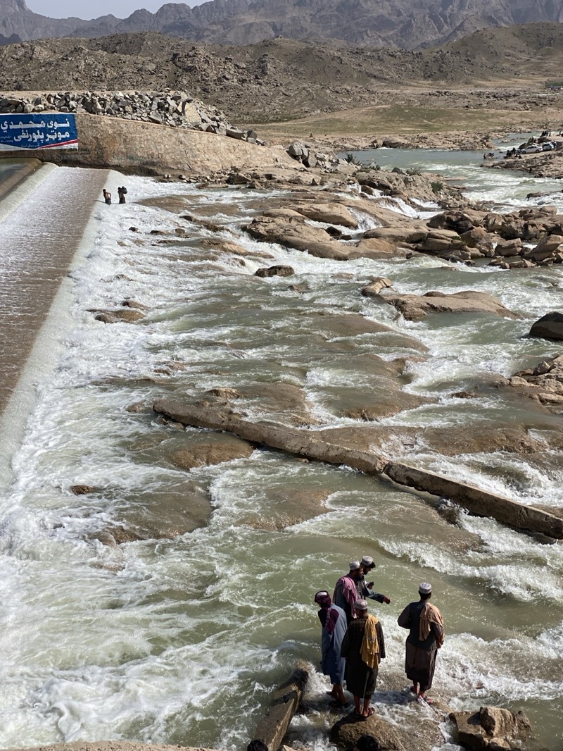 The image depicts a wide, rocky river in Kim, Afghanistan, with rapids flowing downstream. In the foreground, a group of five men wearing traditional Afghan clothing and headwear stand on rocks at the river's edge, observing the water. To the left, a concrete structure spans the river, creating a small waterfall effect. Above it, a sign with text in Arabic is visible. The riverbed is composed of light-colored rocks and boulders. In the background, barren mountains and arid terrain define the landscape. The sky is partly obscured by haze.