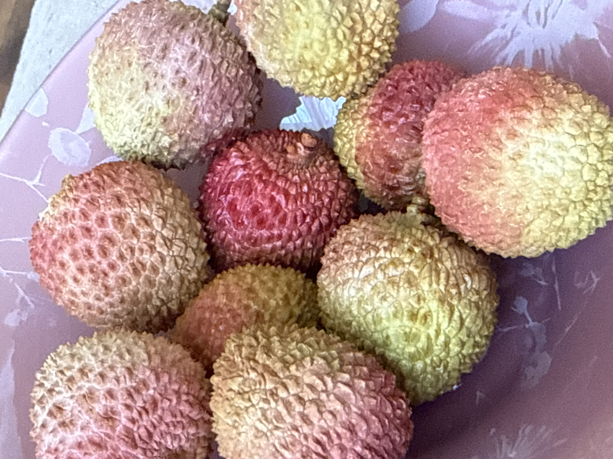 This media file shows a close-up, overhead view of a pile of lychees in a pink bowl. The lychees are round and bumpy, with rough skins that vary in color from pale yellow to pink and red. The bowl has a subtle floral pattern on its surface, visible in the background. There are no people or activities depicted, only the fruit and the dish. The lighting appears to be natural, suggesting it was taken indoors during the daytime. No text is visible in the image. The context provided suggests this image might have been taken in Chelyabinsk, Russia, though there are no visual cues to confirm this specific location.