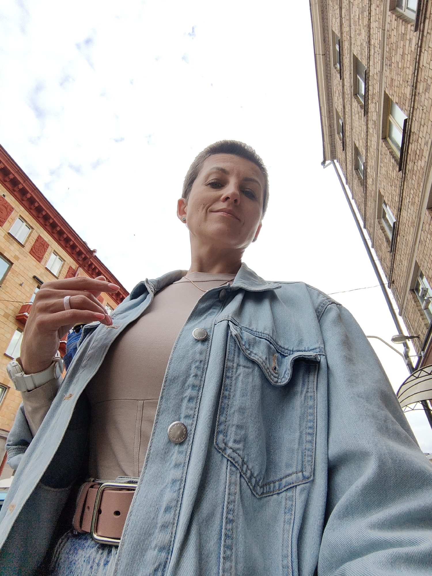 A medium shot, low-angle selfie of a woman with short, dark hair and short bangs is taken outdoors, with two buildings and the sky visible. The woman is wearing a light beige shirt, a light blue denim jacket, and a light pink belt. Her left hand, adorned with a ring, is raised towards her face. She is looking directly at the camera. The building on the left is red with white-framed windows and red decorative accents. The building on the right is made of brown brick with several windows. The sky is bright and overcast. This image was taken in Minsk, Belarus.