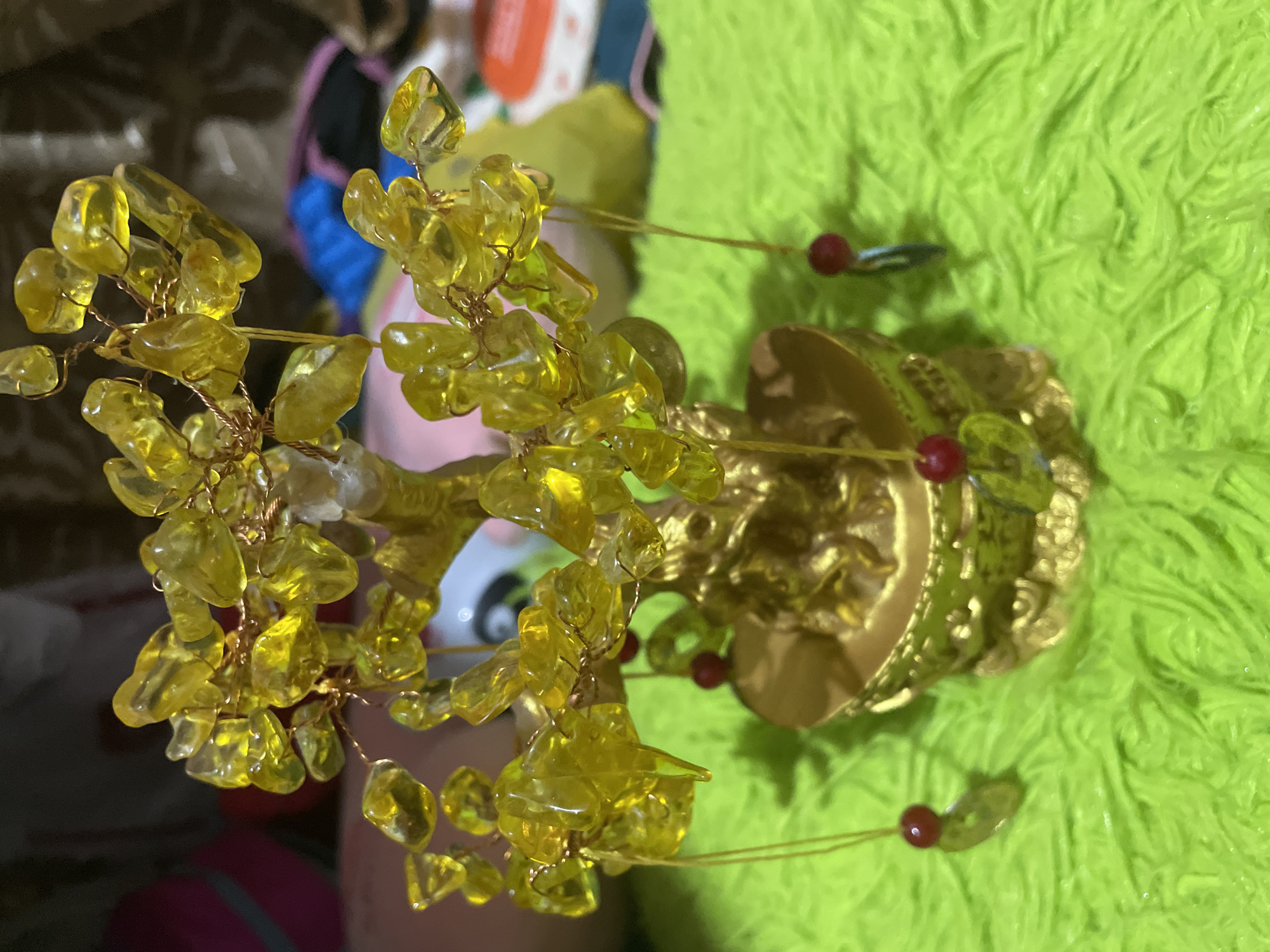 A close-up, slightly rotated image focuses on a small, golden-colored, tree-like ornamental object with yellow crystal-like leaves. The base of the "tree" is elaborate, resembling a miniature golden temple or shrine. The background is a mix of textures and colors, including a bright green, soft-looking material that could be fabric or a plush surface. Additional blurred objects are visible in the background, including a pink surface, a multi-colored pattern, and brown tones. There is no evidence of Davao City, Philippines. No people, actions, or interactions are visible.