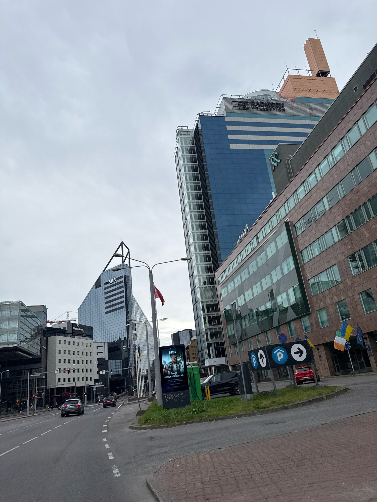 The image shows a street scene in Tallinn, Estonia, with modern skyscrapers dominating the skyline. The perspective is from a street corner, looking up at several tall buildings. One prominent building features the "SEB" logo and has a distinctive triangular shape. Another building, labeled "Radisson," has a distinctive architectural element at its top.
In the foreground, a street with cars is visible, including a silver sedan and a red car. There are traffic lights and road markings. On the right side of the street, a sidewalk area with paving stones and grass can be seen. A sign with parking symbols and directional arrows is present. Several flags are displayed on the building to the right, including the Ukrainian flag, an Irish flag, and the European Union flag.
A digital advertisement billboard is also visible, showing an image of a man and text in Estonian. The sky is overcast, suggesting a cloudy day. The overall impression is of a modern, commercial district within the city of Tallinn.