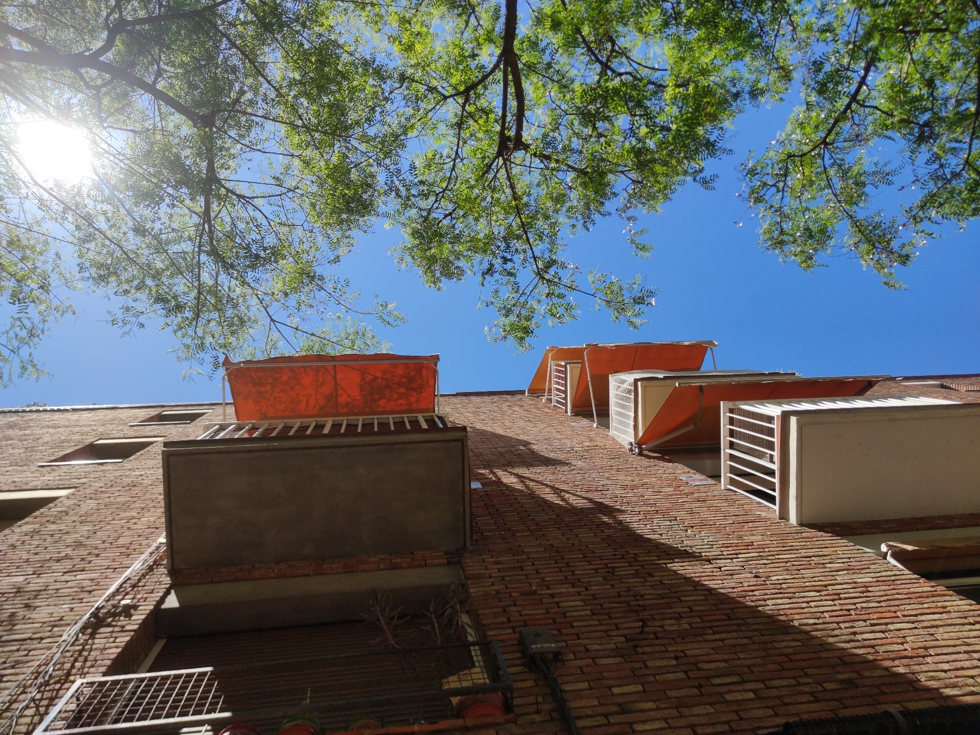 The image shows a low-angle view of a brick building under a clear blue sky, partially obscured by tree branches and leaves. The sun is visible in the upper left corner, creating lens flare. The building has projecting balconies or structures, some with orange awnings or sunshades. There are also some visible utility fixtures on the lower part of the brick wall. No people or animals are visible in the image. The location is Unknown, Unknown.