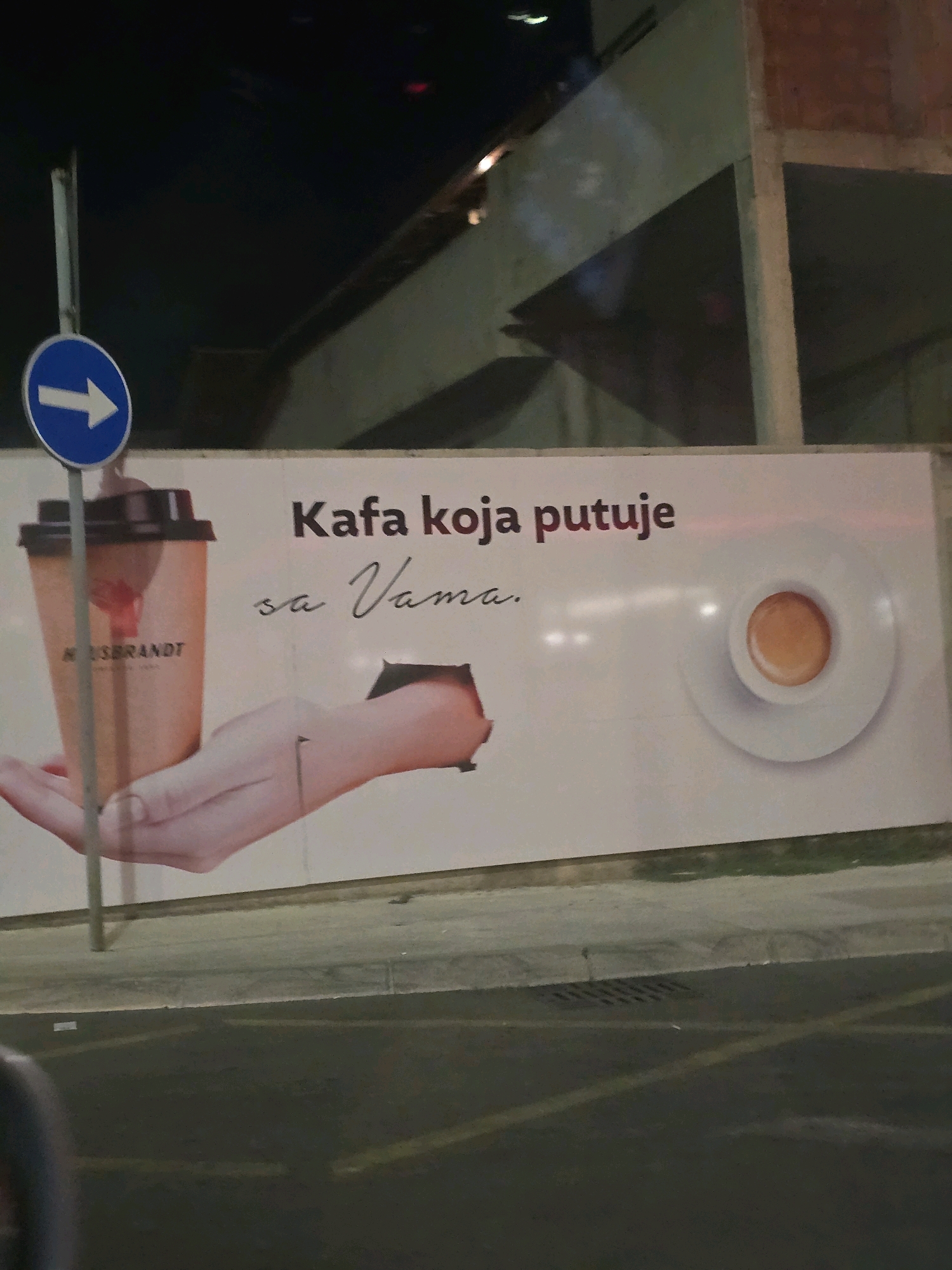 The image shows a billboard in Podgorica, Montenegro, advertising Hausbrandt coffee. The billboard features a large image of a Hausbrandt coffee cup held by a hand that appears to be emerging from a cut-out section of the billboard. The text on the billboard reads "Kafa koja putuje sa Vama", which translates to "Coffee that travels with you". To the left of the billboard, a blue directional sign with a white arrow pointing to the right is visible. The billboard is illuminated, and the surrounding area appears to be a street or sidewalk at night.
