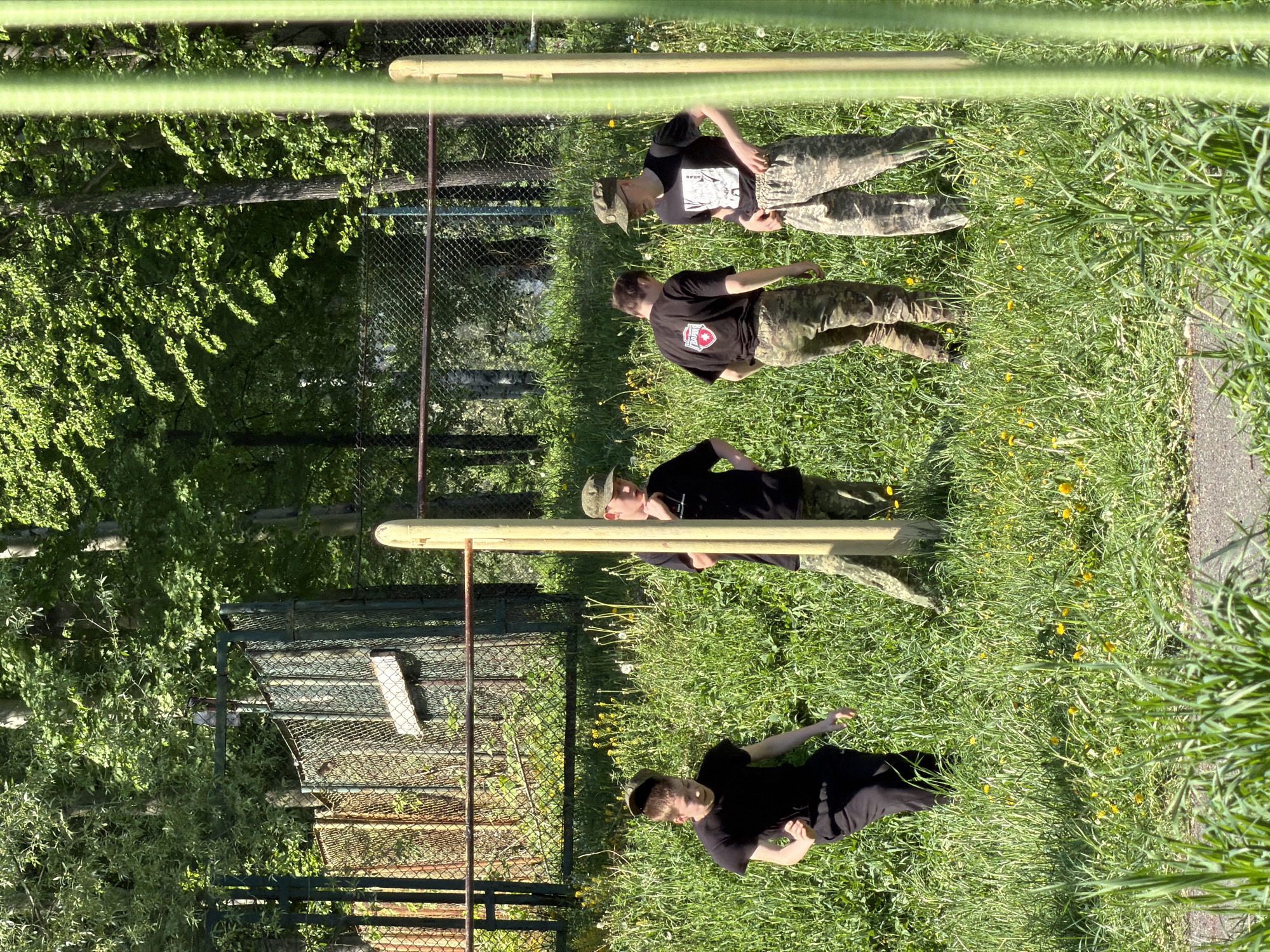 The image depicts a scene in a grassy area, likely a park or training ground, with a wooden obstacle course. Four individuals are present. Three are wearing camouflage pants and black shirts, while the fourth wears all black. One is wearing a camouflage hat. The individuals appear to be either taking a break or awaiting instructions.
In the background, a chain-link fence borders a wooded area. The location is consistent with the city of Oreshniki, Russia, based on the vegetation and infrastructure visible in the image, although the image does not provide a direct location reference. The time of day appears to be midday, based on the lighting and shadows.