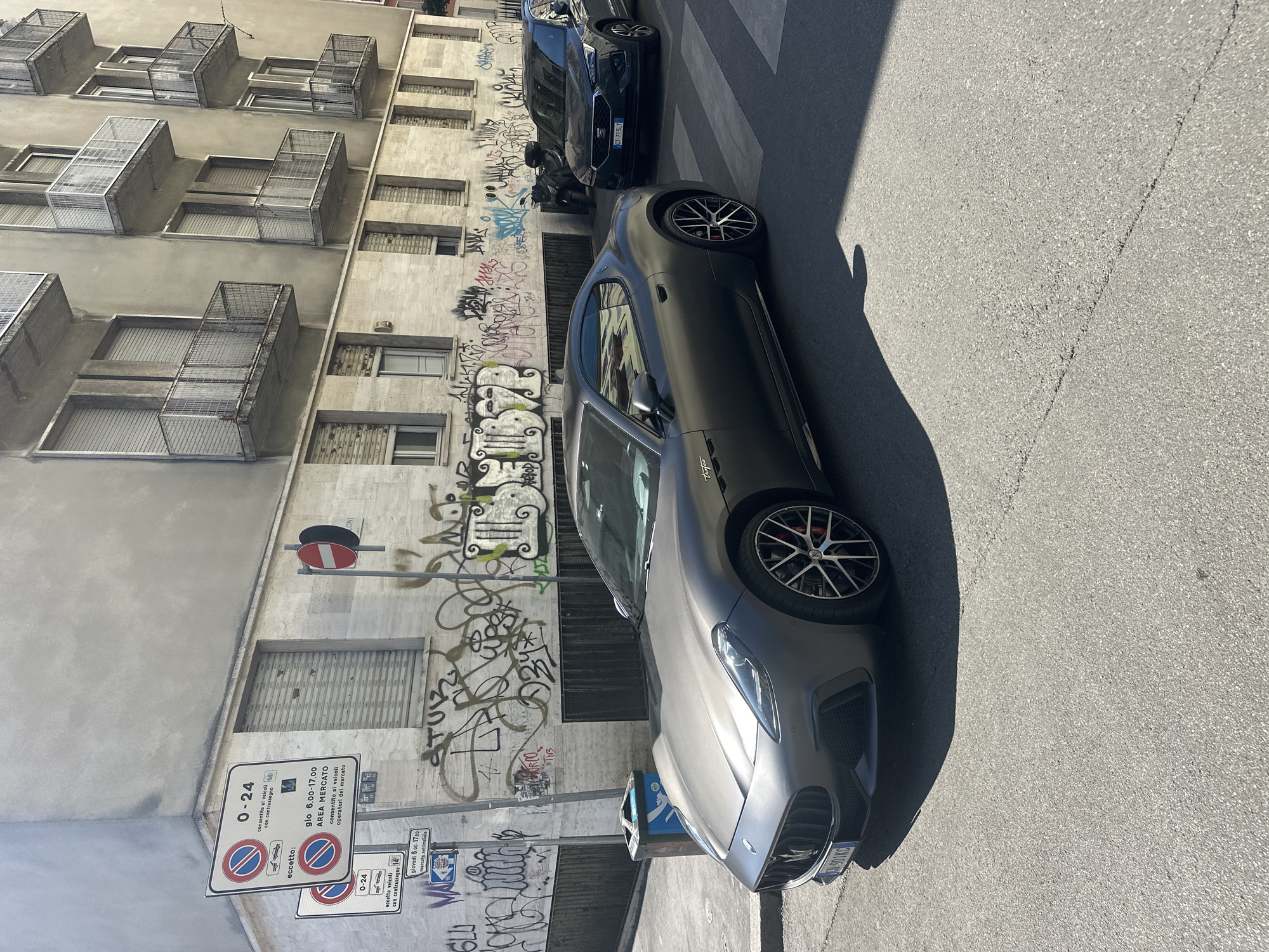 The image shows a gray Maserati parked on the side of a street in Milan, Italy. The building behind the car is covered in graffiti, and there are several signs posted on the building, including one that says "AREA MERCATO." Another sign says "0-24." A black car is parked behind the Maserati and a crosswalk is visible to the right of the frame. The building has metal caging around some of the windows.