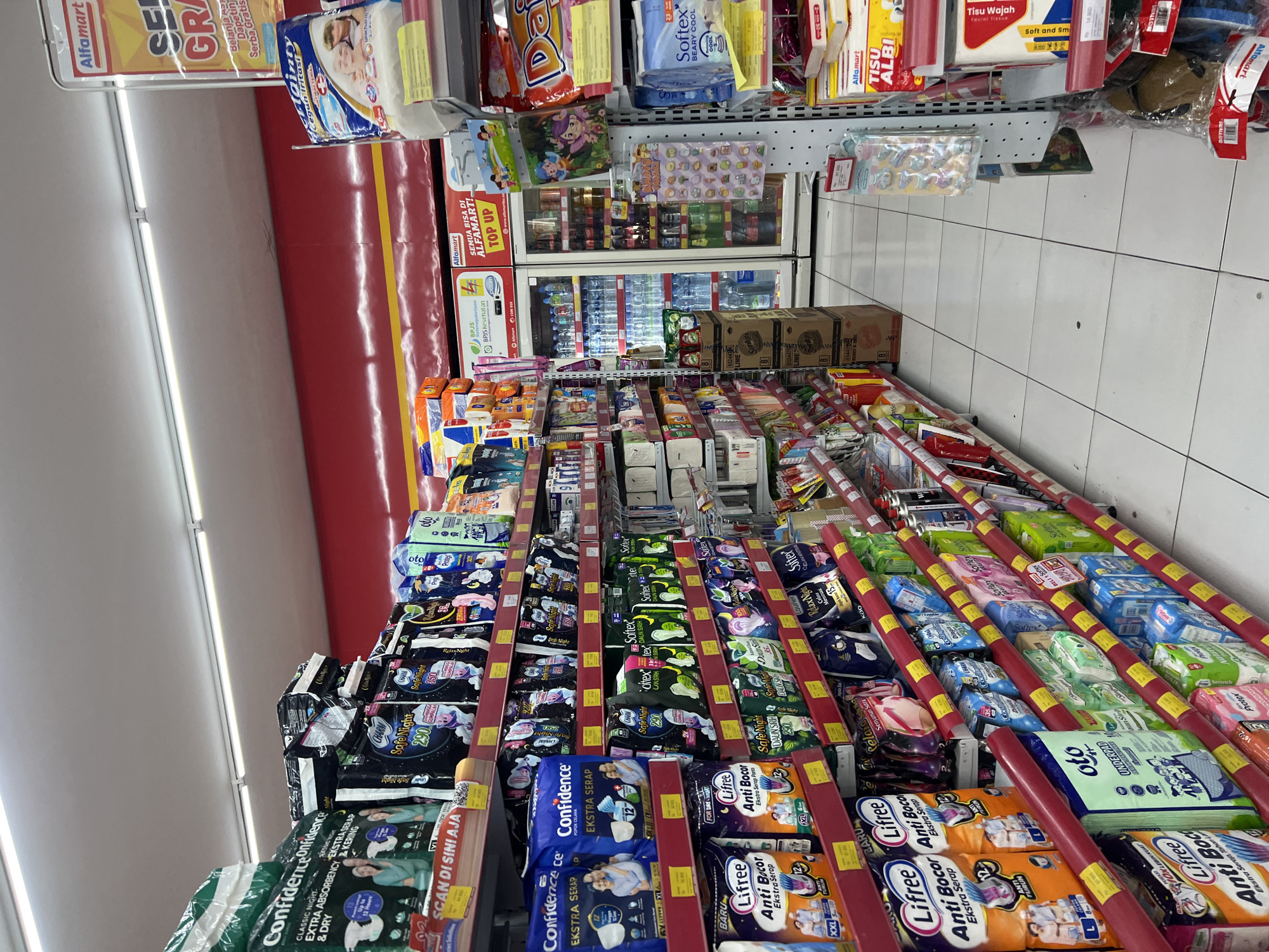 This is a photograph taken inside an Alfamart convenience store, likely in Kuta, Indonesia, given the signage and product packaging. The perspective is from a low angle, looking down an aisle filled with shelves stocked with various products, predominantly adult diapers and other personal hygiene items.
The shelves are made of metal with red support bars and yellow price tag holders. The products are arranged neatly, with multiple brands and types of diapers visible, including "Confidence," "Charm SafeNight," "Lifree Anti Bocor," and "Sofftex." Many packages are colorful and feature images of babies or families. There are also other items such as bottled water, snacks, and tissues on display.
In the background, two glass-doored refrigerators are visible, stocked with drinks. The store has a bright, fluorescent-lit ceiling and tiled flooring. Signs advertising promotions, such as "SEMBEL SERBA GRATIS" (Free Everything) and "TOP UP," are displayed. There are no people clearly visible in the immediate foreground or performing any actions. The overall scene depicts a well-organized retail environment typical of a convenience store.