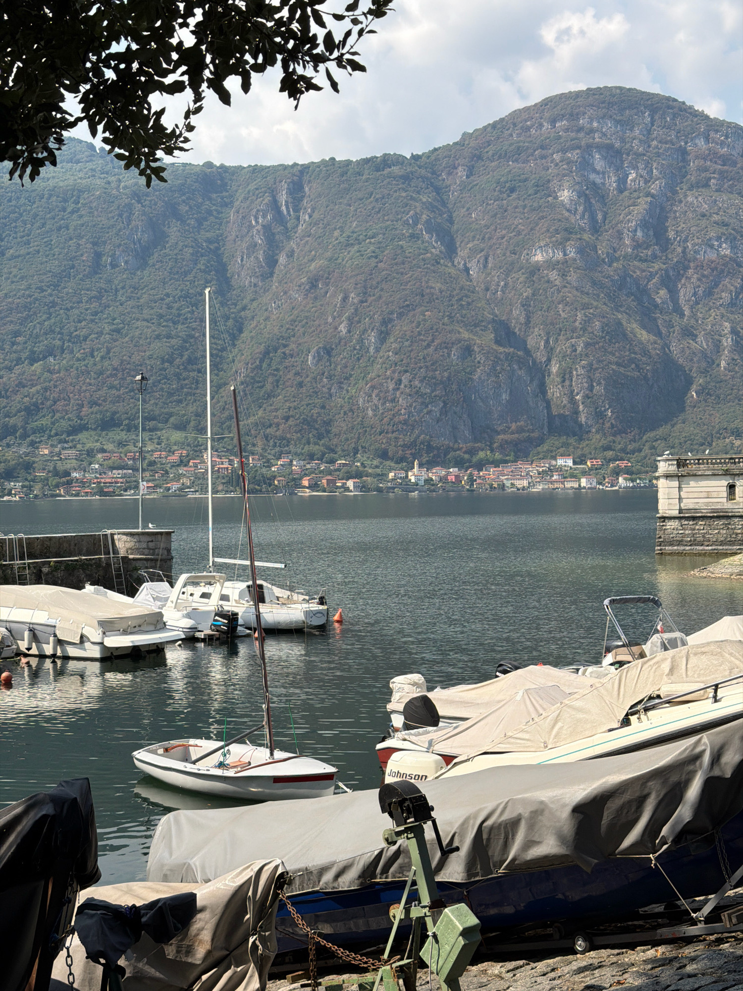 In Mandello del Lario, Italy, a still lake is bordered by a lush green mountainside. The lake hosts several boats docked at a quay. In the foreground, various boats are covered with tarpaulins, suggesting they are either stored or undergoing maintenance. A small white sailboat with a single mast floats in the water. Further back, several larger white motorboats are also docked. The town of Mandello del Lario is visible on the distant shore, with colorful buildings dotting the hillside. A stone structure with arches stands on the right side of the frame, overlooking the lake. The sky is partly cloudy, casting soft light on the scene. No people are visible in the image.