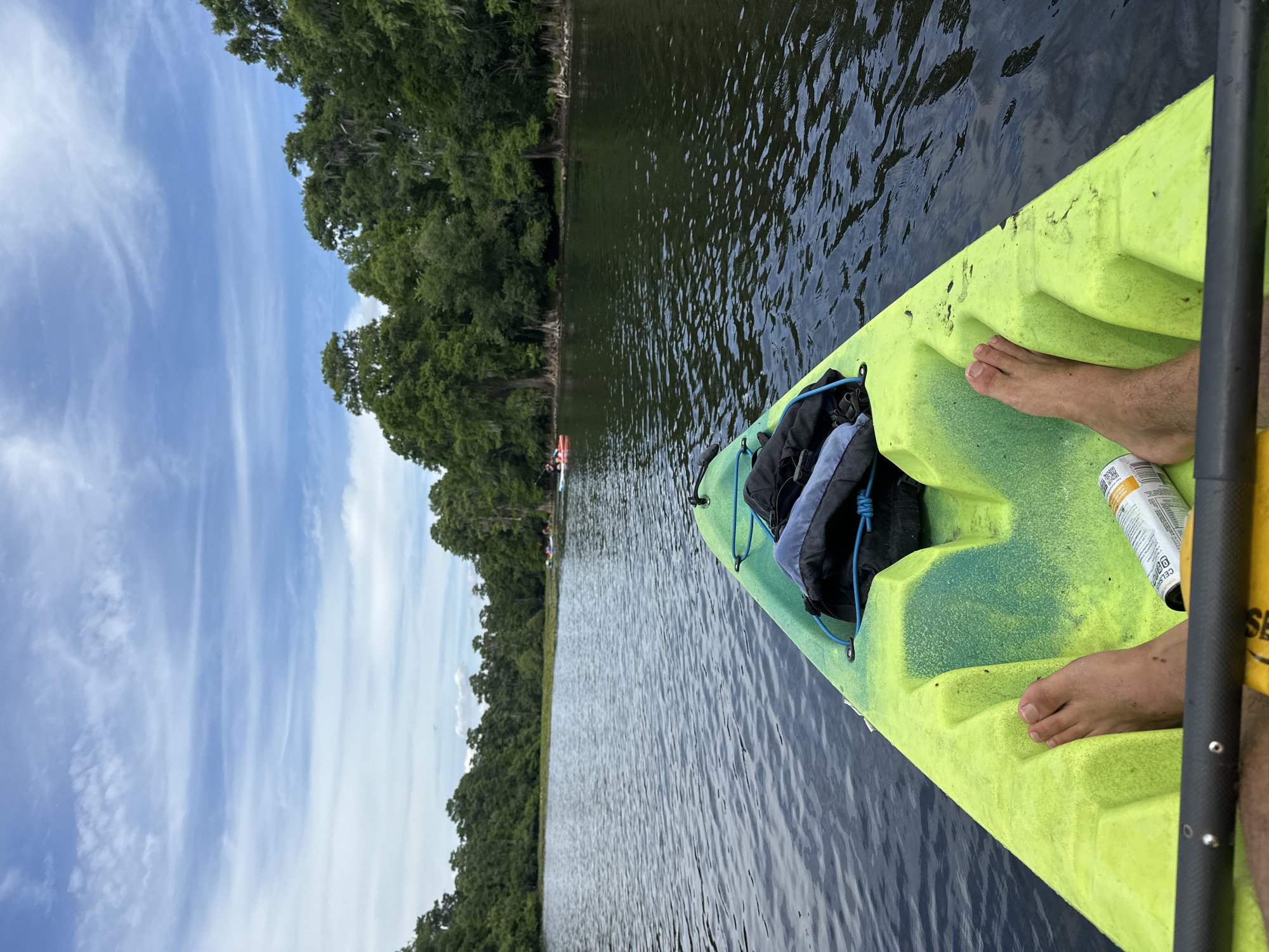 The image captures a first-person perspective from a bright neon green and yellow kayak on a calm body of water, likely a river or lake. The kayaker's bare feet rest on the kayak's textured surface, with a black paddle shaft partially visible on the right. A silver can, possibly bug spray or a beverage, is positioned near the feet, displaying the partial text "CELSIUS" and other small, illegible words. A black dry bag or backpack is secured to the front of the kayak.
The scene unfolds under a broad, light blue sky adorned with wispy white clouds, indicating fair weather. Along the left side of the water, a dense and lush green treeline stretches into the distance, with some trees showing Spanish moss, common in humid environments like Florida. Further down the waterway, faint outlines of other kayaks or small boats can be observed. The water itself is dark and reflects the sky and trees, with gentle ripples disturbing its surface.
The activity depicted is kayaking, suggesting a peaceful outdoor excursion. The time of day appears to be daytime, with clear natural lighting. The overall impression is one of relaxation and connection with nature, consistent with exploring the scenic waterways near Tampa, United States.